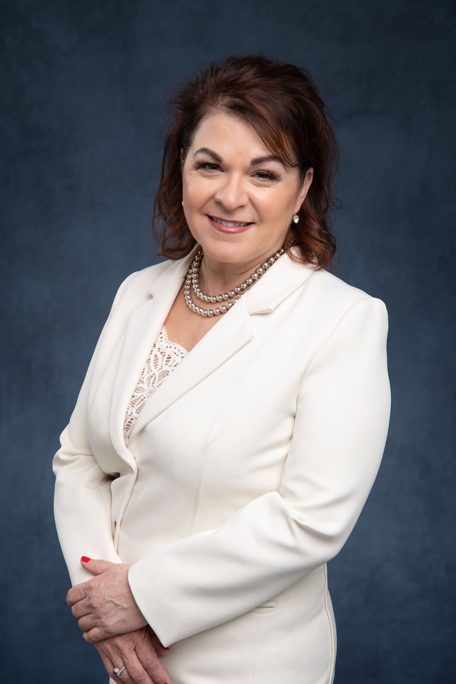Professional studio portrait of someone wearing a white blazer and pearl necklace against a dark background.