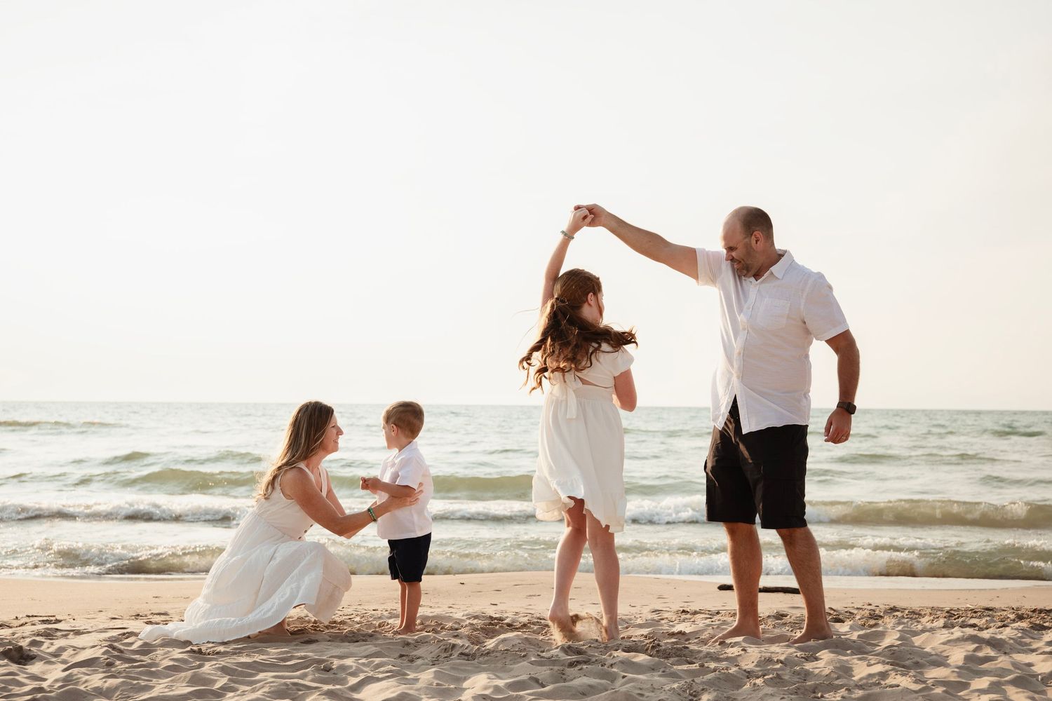 Family dressed in white enjoying a playful moment together on a sunny beach by the ocean at sunset.