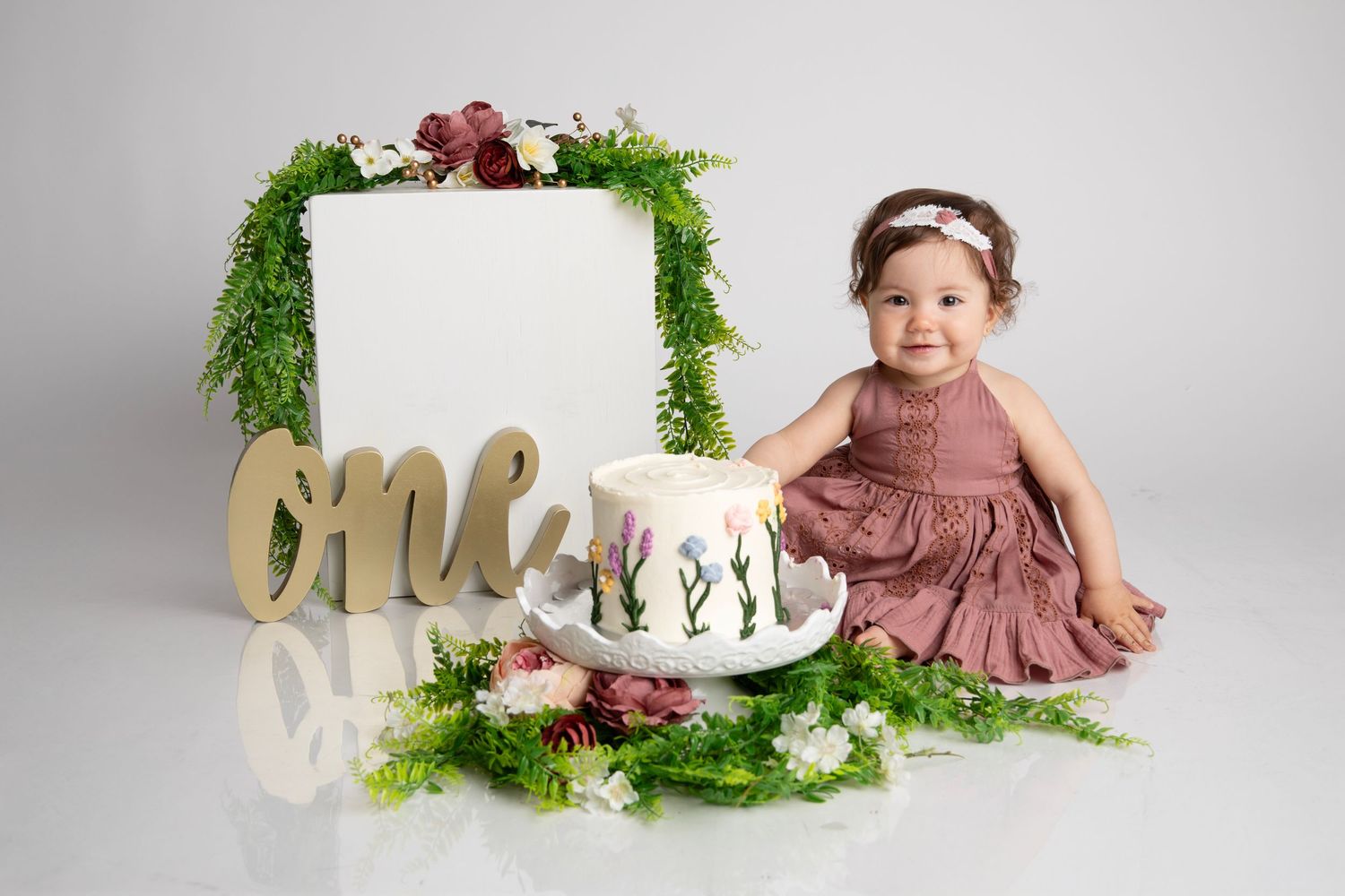 Studio portraits with greenery and floral decorations showing baby in mauve dress with a custom cake for her 1st birthday photos.