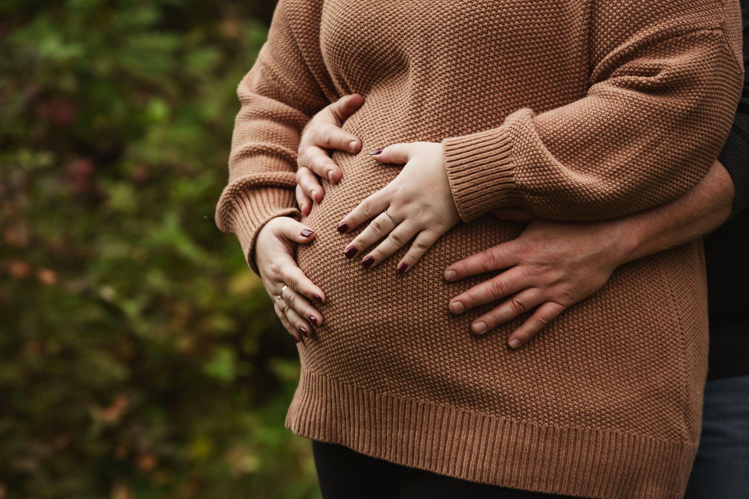 Close up of hands cradling pregnant belly in brown sweater against forest background.
