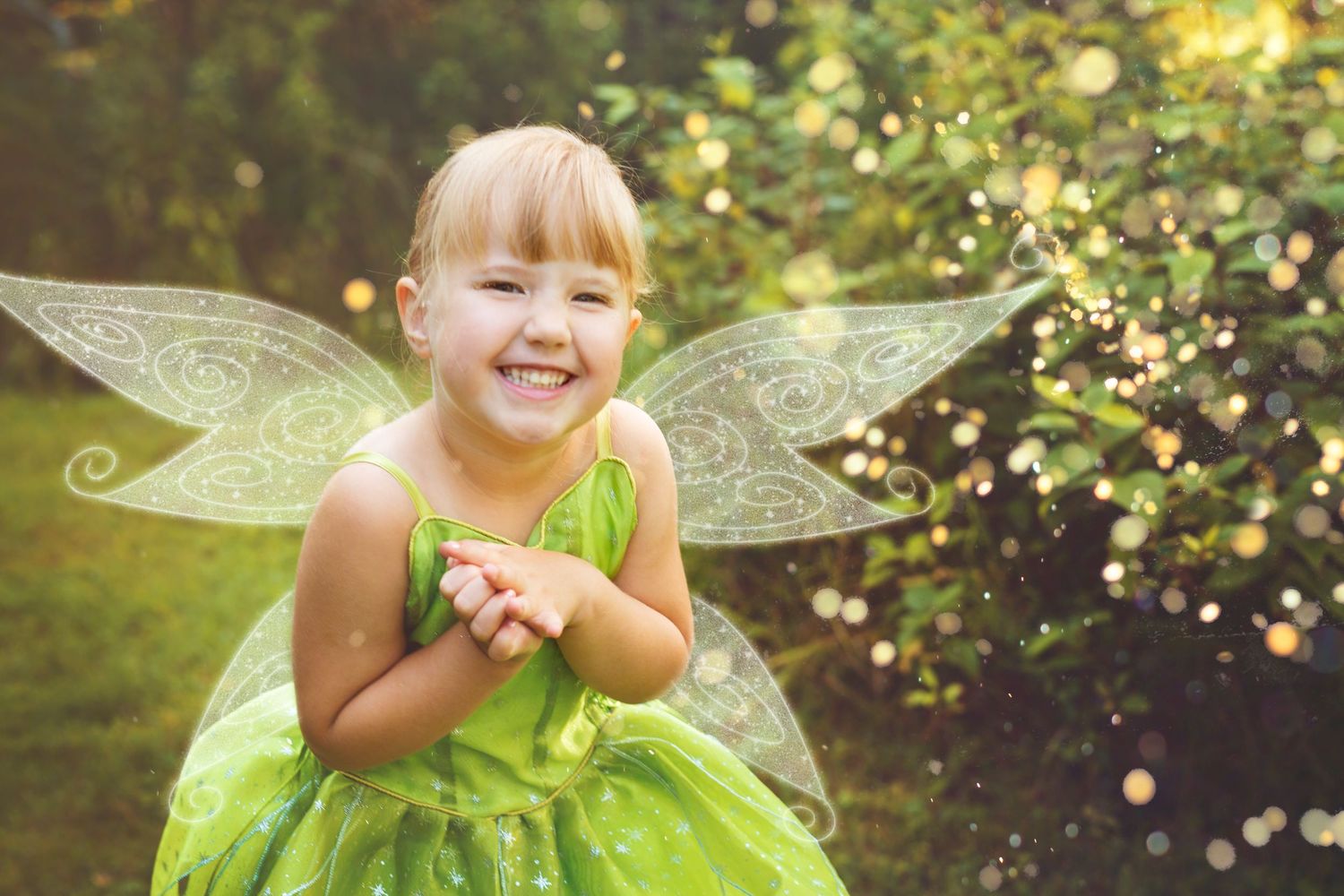 Young child wearing green fairy costume with wings laughs joyfully in sunlit garden setting.