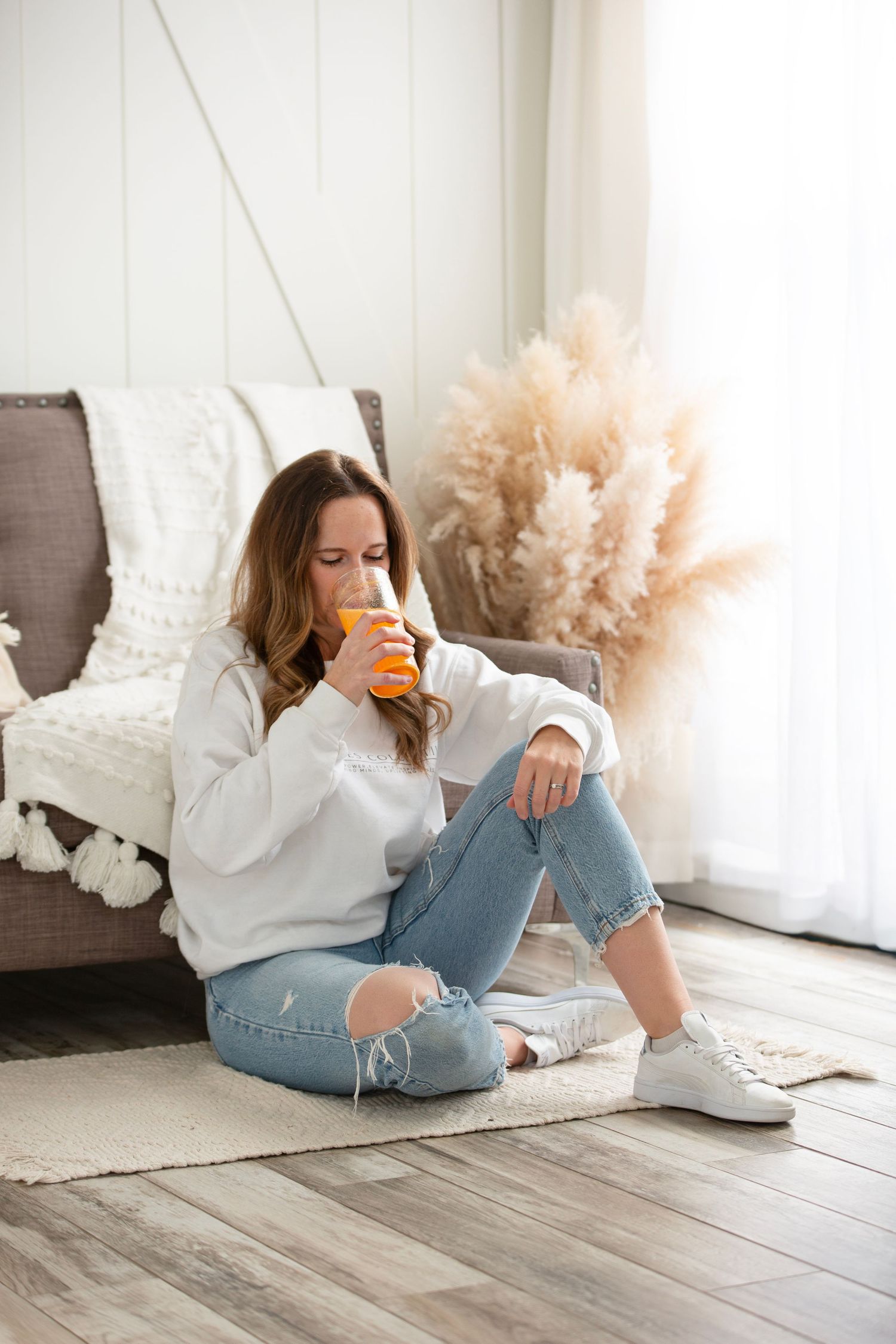 Person in white sweater and ripped jeans relaxing on floor cushion with orange drink near pampas grass decor.