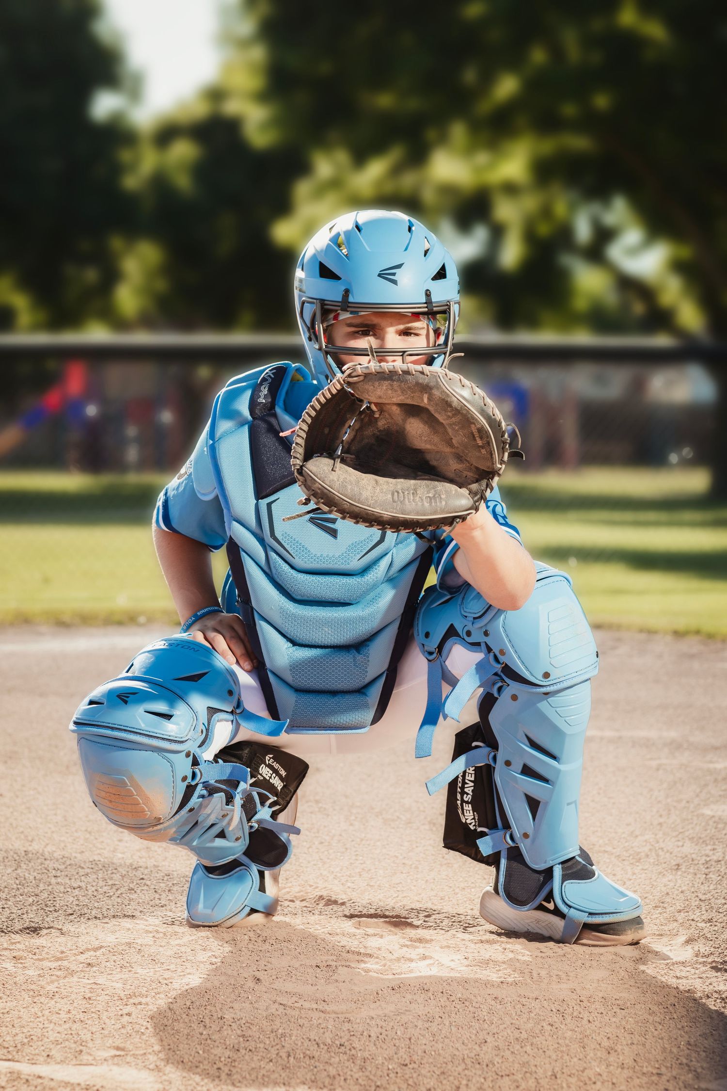 Young baseball catcher in full blue Easton gear crouching at home plate, ready to receive a pitch on a sunny day.