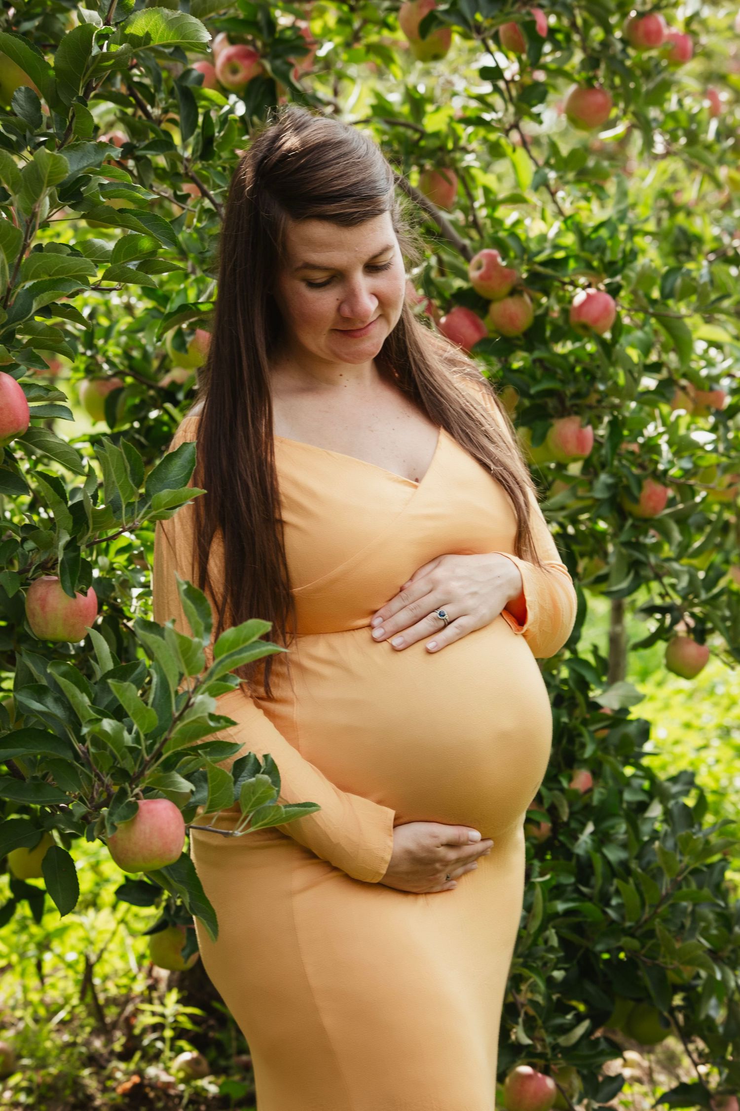 Maternity photo of woman in yellow dress among apple trees in orchard.