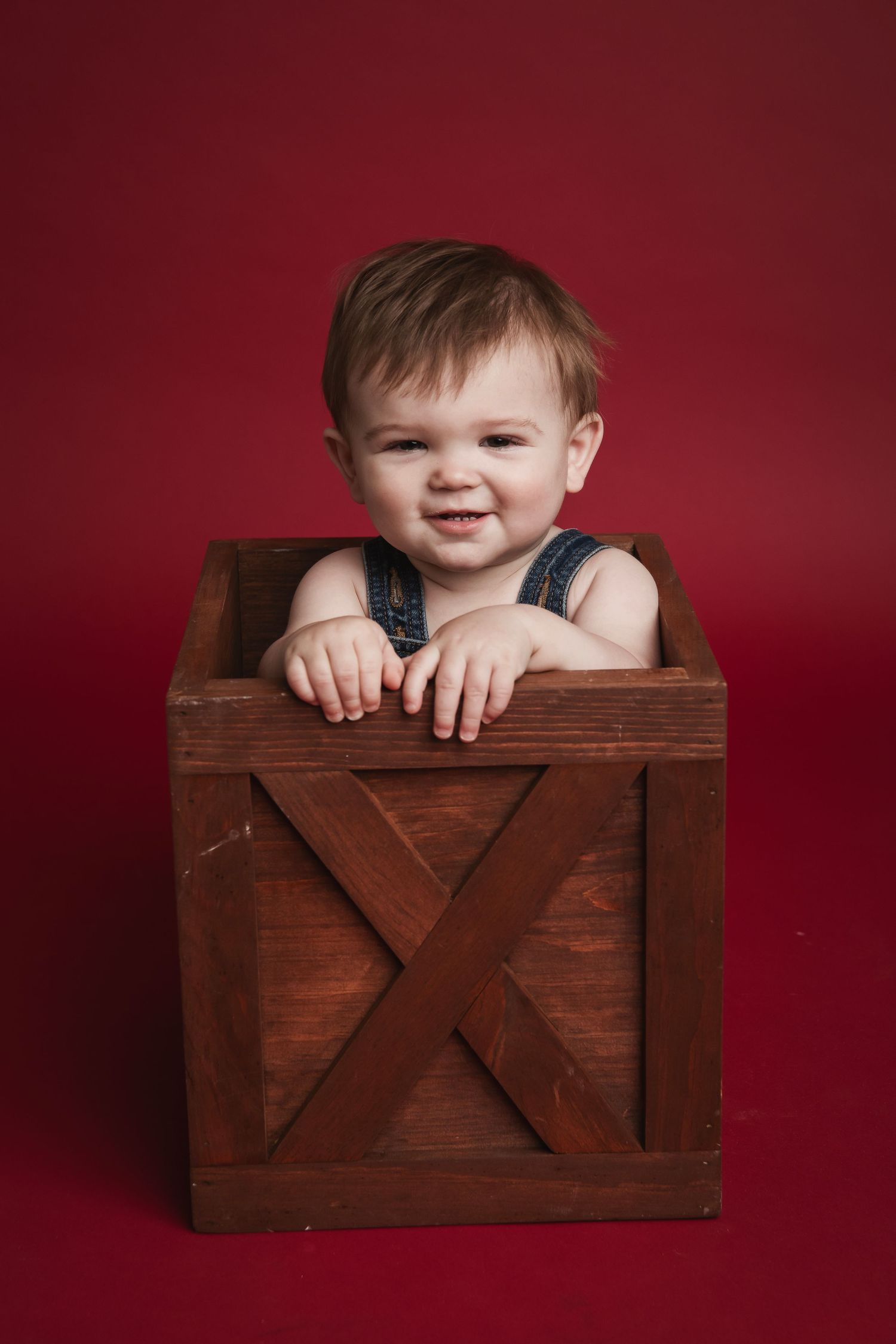 A cheerful toddler sits inside a rustic wooden crate against a burgundy backdrop.