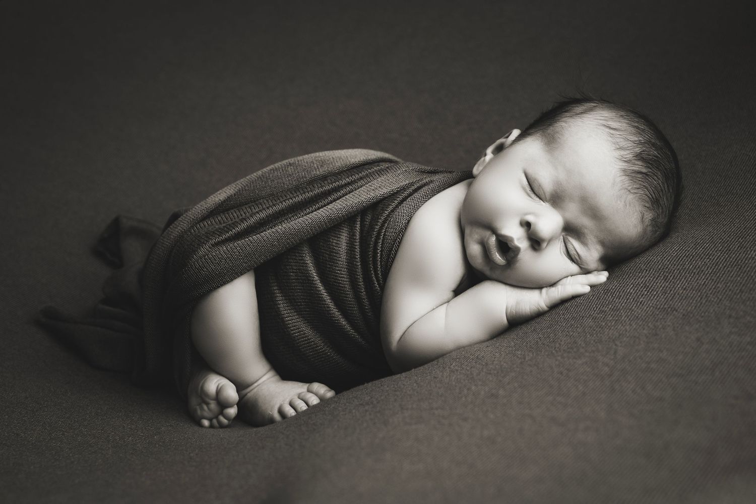Sleeping newborn wrapped in dark fabric on black background in dramatic lighting.