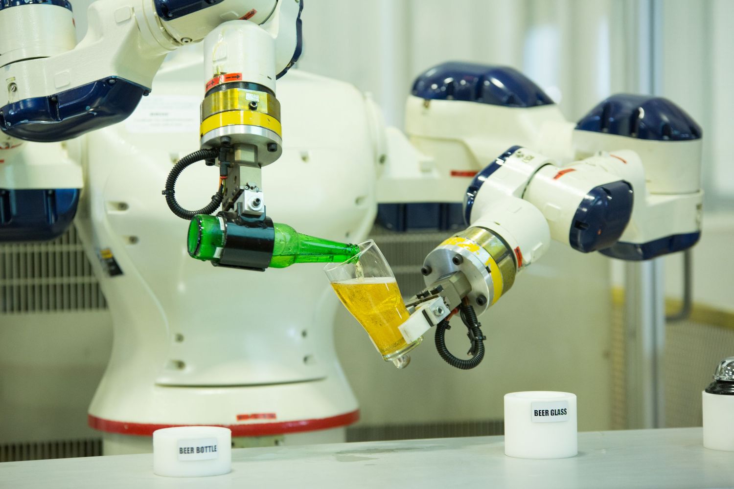 Robotic arms pouring yellow liquid between beakers in a laboratory testing environment.