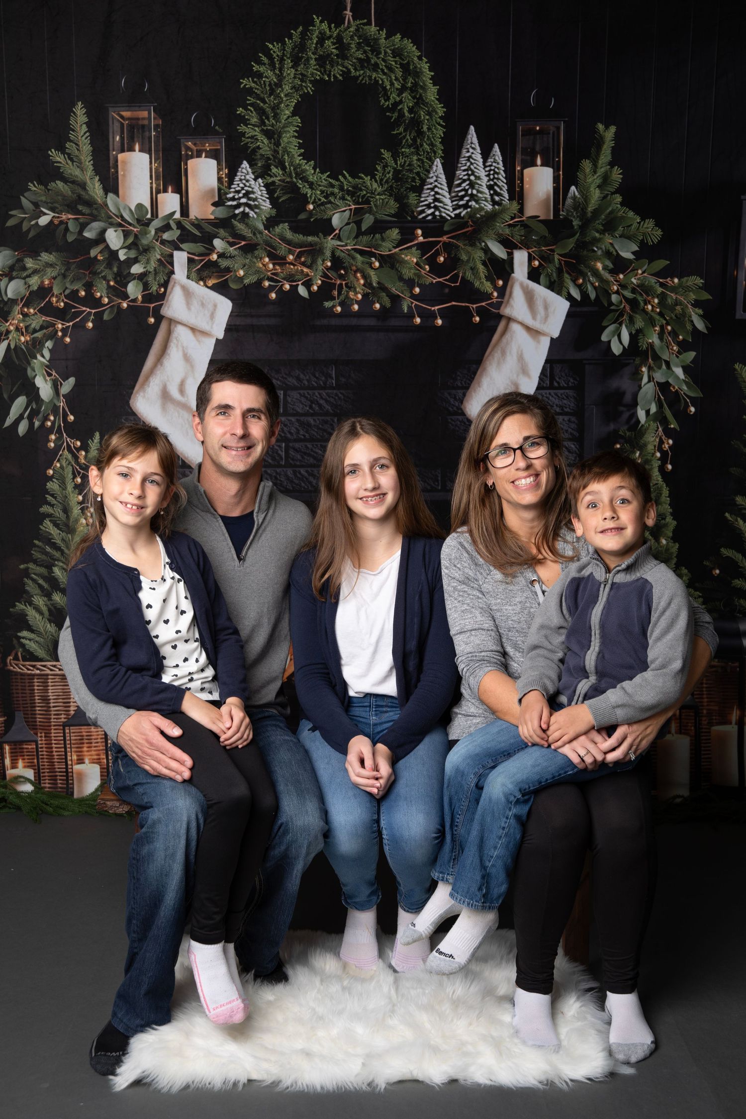 Family wearing coordinated blue jeans and casual tops pose together on white fur rug for holiday photos.