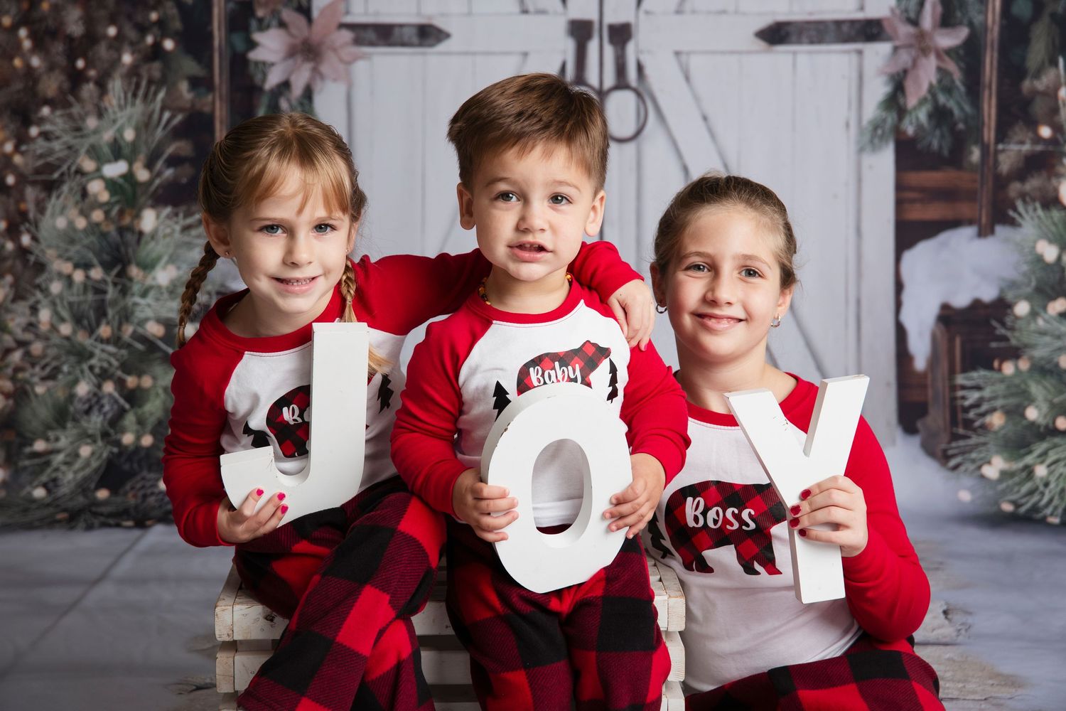 Three people in red and white Christmas outfits pose with JOY letters in front of a decorated tree.