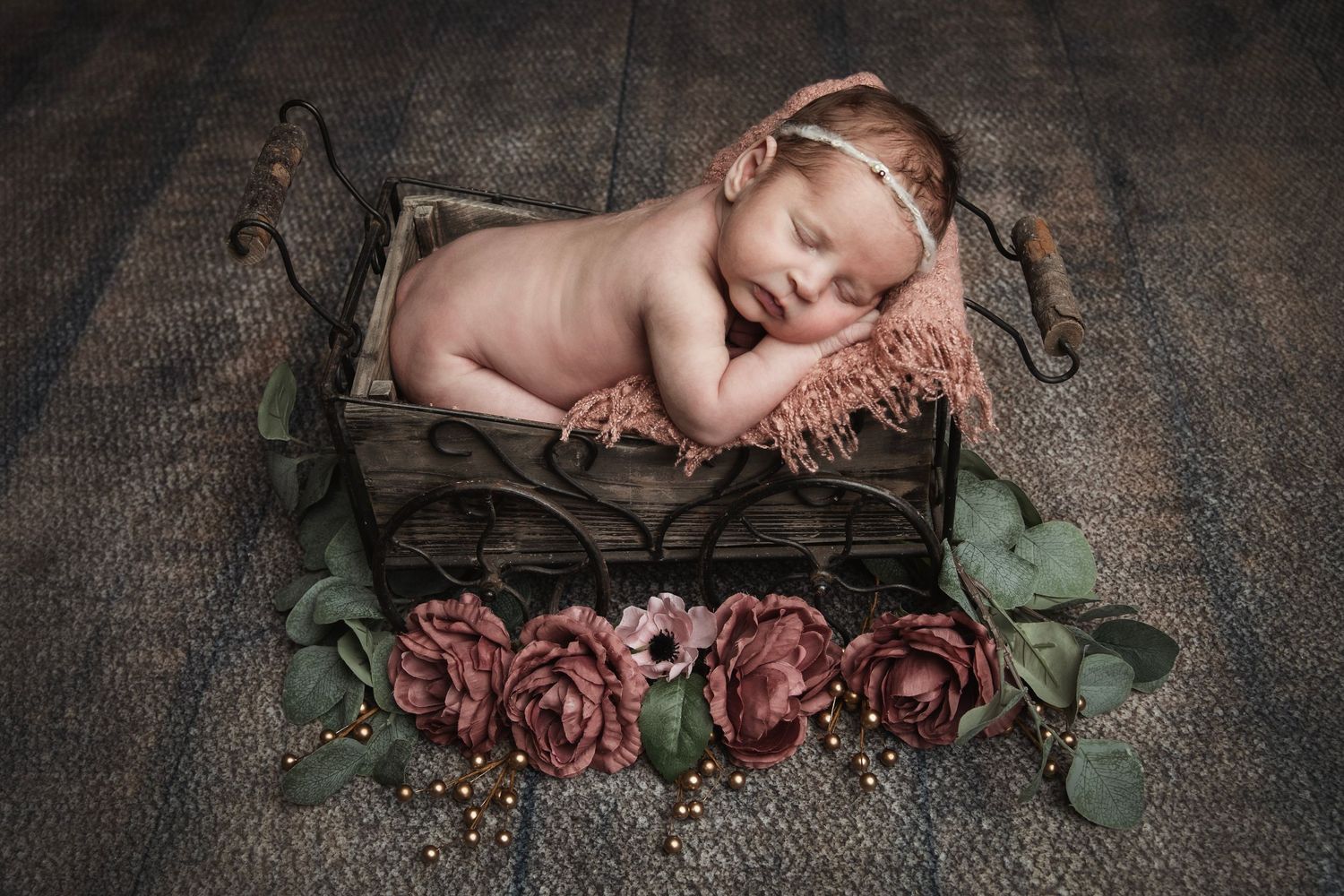 Sleeping newborn in rustic wooden box surrounded by deep red roses and greenery.