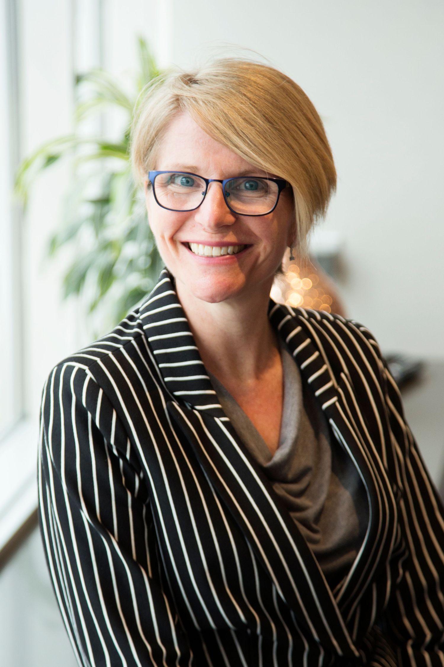 Professional wearing black and white striped blazer, glasses and gray scarf smiles warmly in office setting with plant.