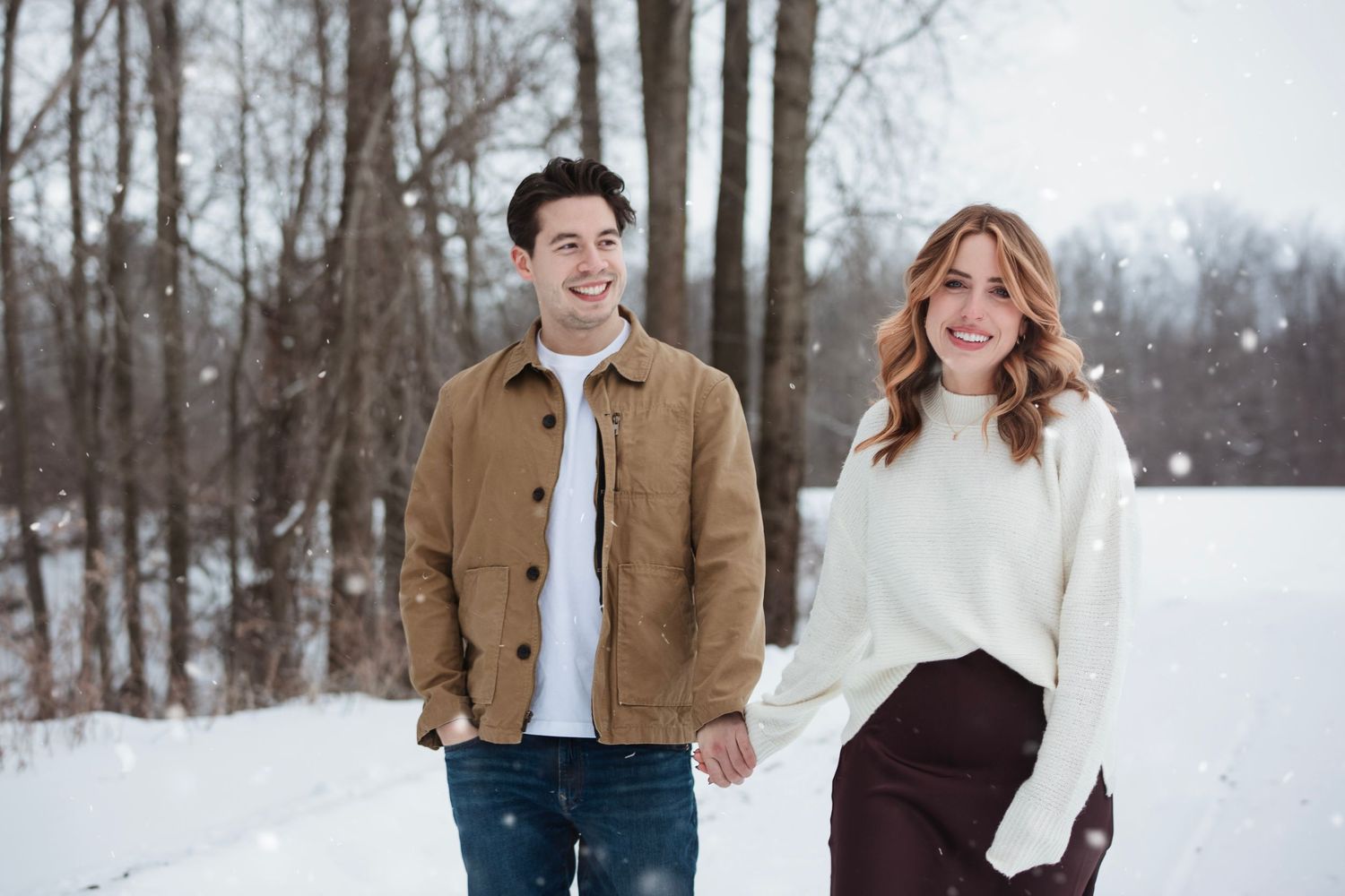 A couple walks hand in hand through a snowy winter forest landscape wearing warm coats and smiling.