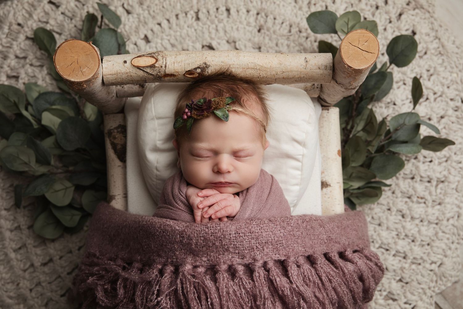 A cozy newborn photo collage with baby wrapped in mauve knit blankets surrounded by eucalyptus greenery and wooden accents.