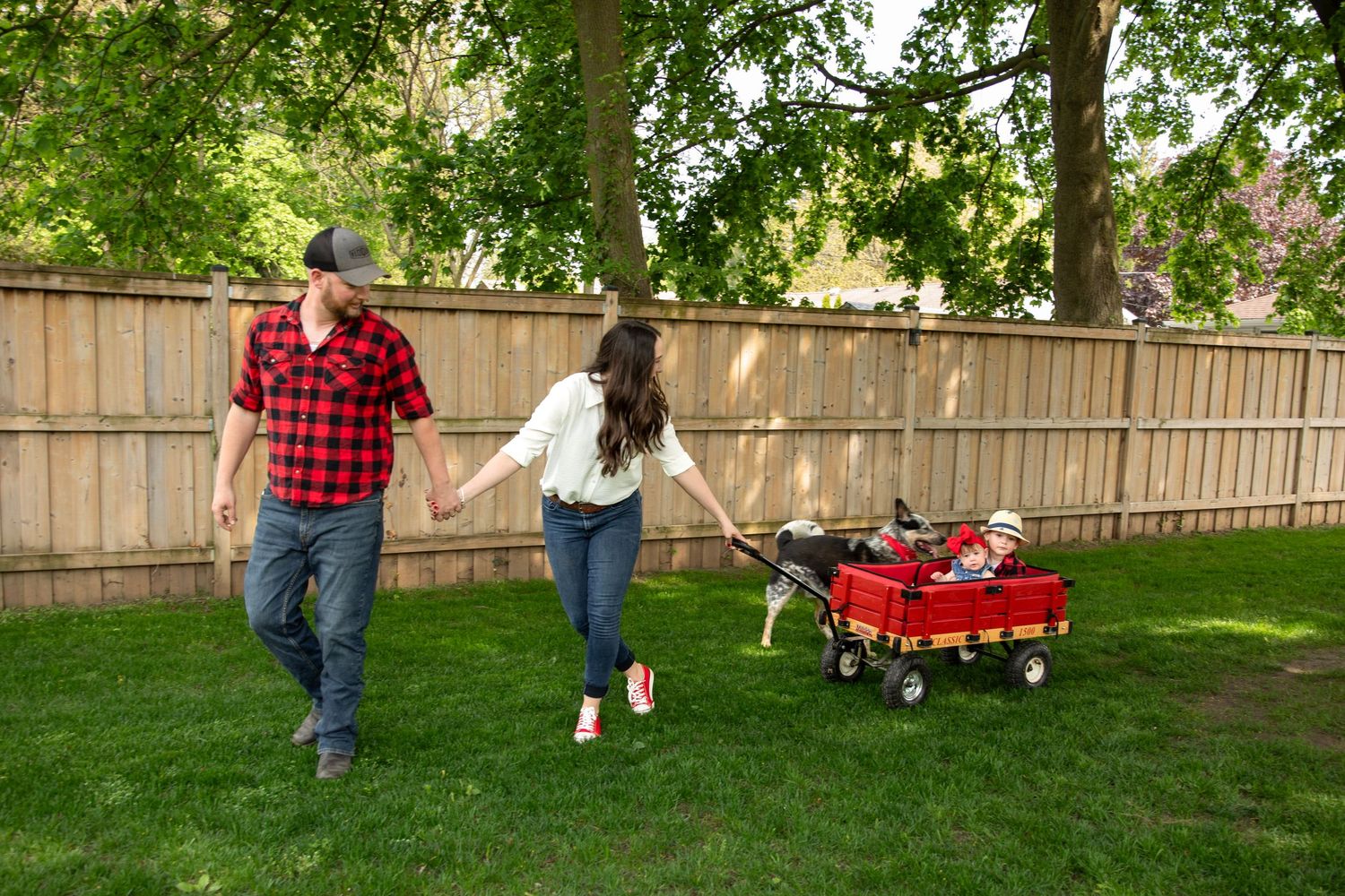 A couple holds hands while pulling a red wagon with a child through a lush green backyard with a wooden fence.