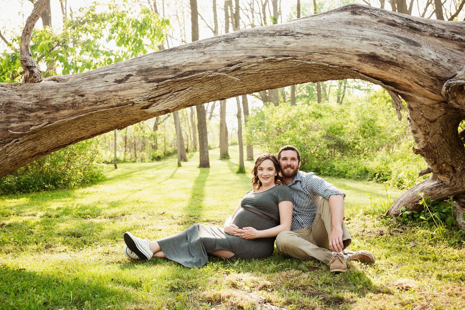 Maternity photo session with couple sitting on fallen tree in sunlit forest.