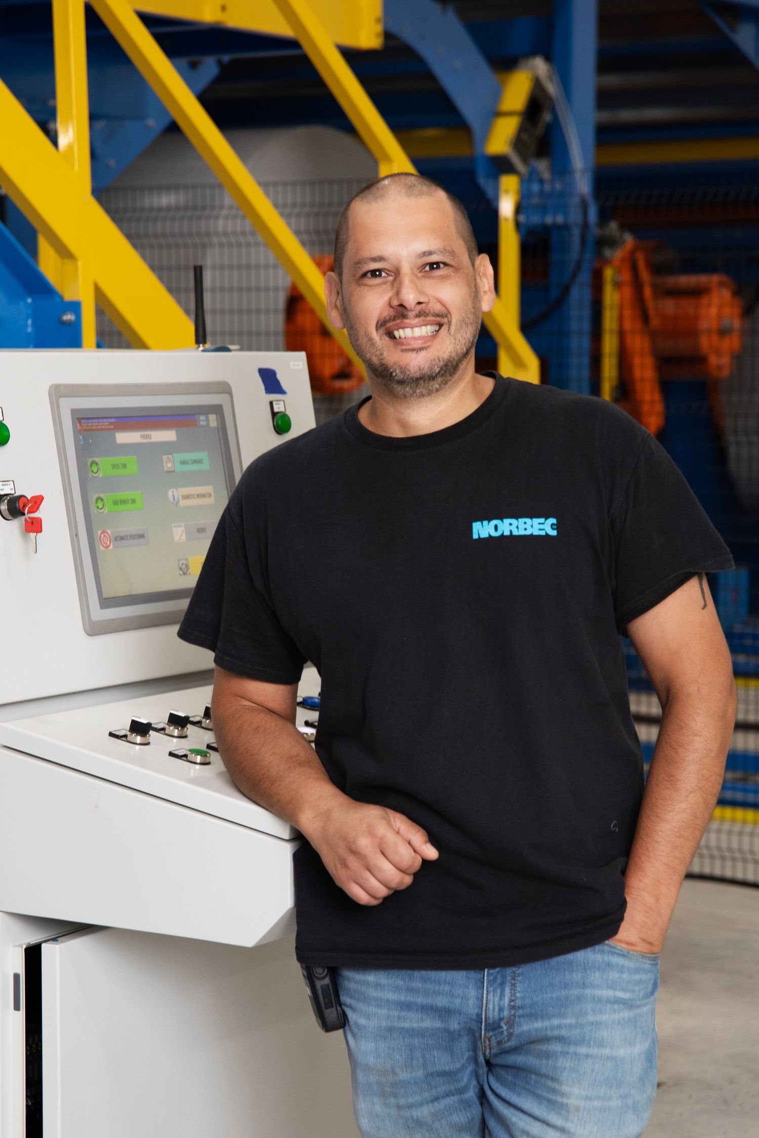Manufacturing employee in Norbec branded black t-shirt standing by industrial control panel in warehouse setting.