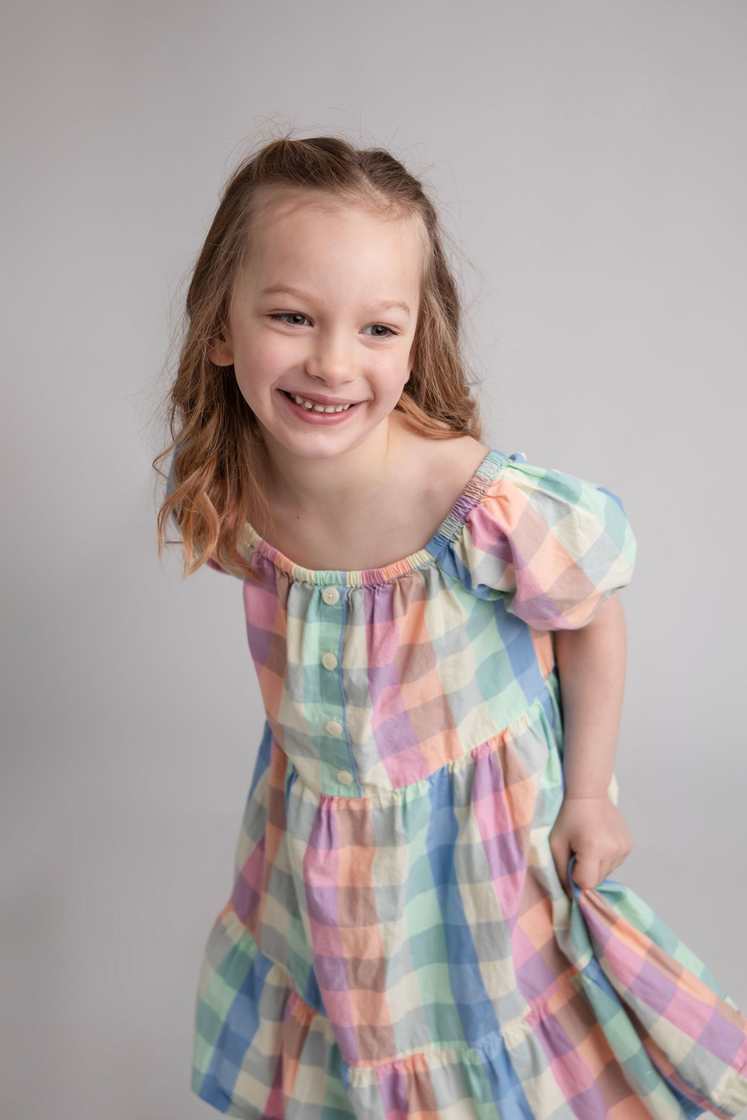 Young girl in pastel plaid dress smiling against grey background during portrait session.