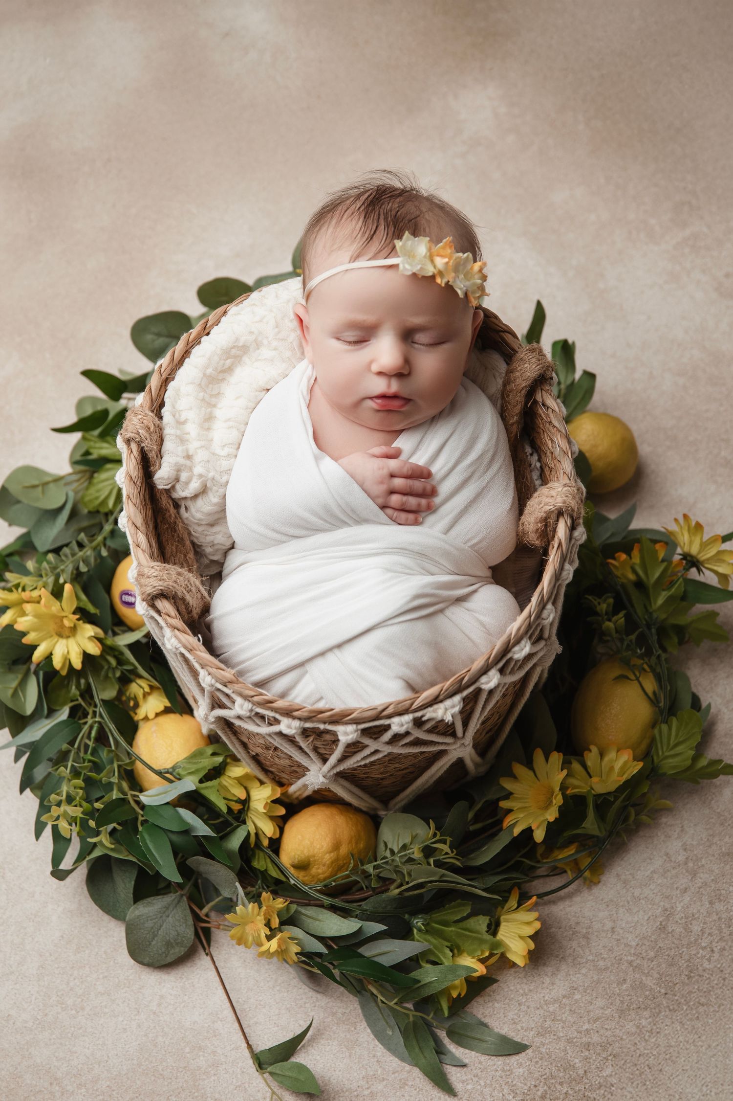 Sleeping newborn wrapped in white swaddle nestled in basket with lemons and sunflowers.