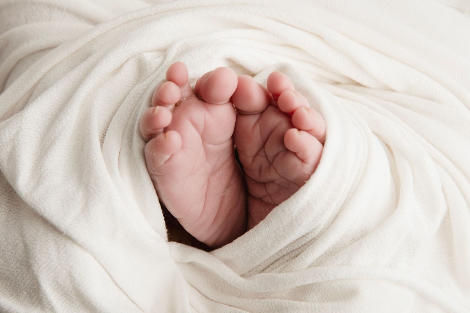 Tiny newborn hands peek out from soft white blankets in an artistic close-up photo.