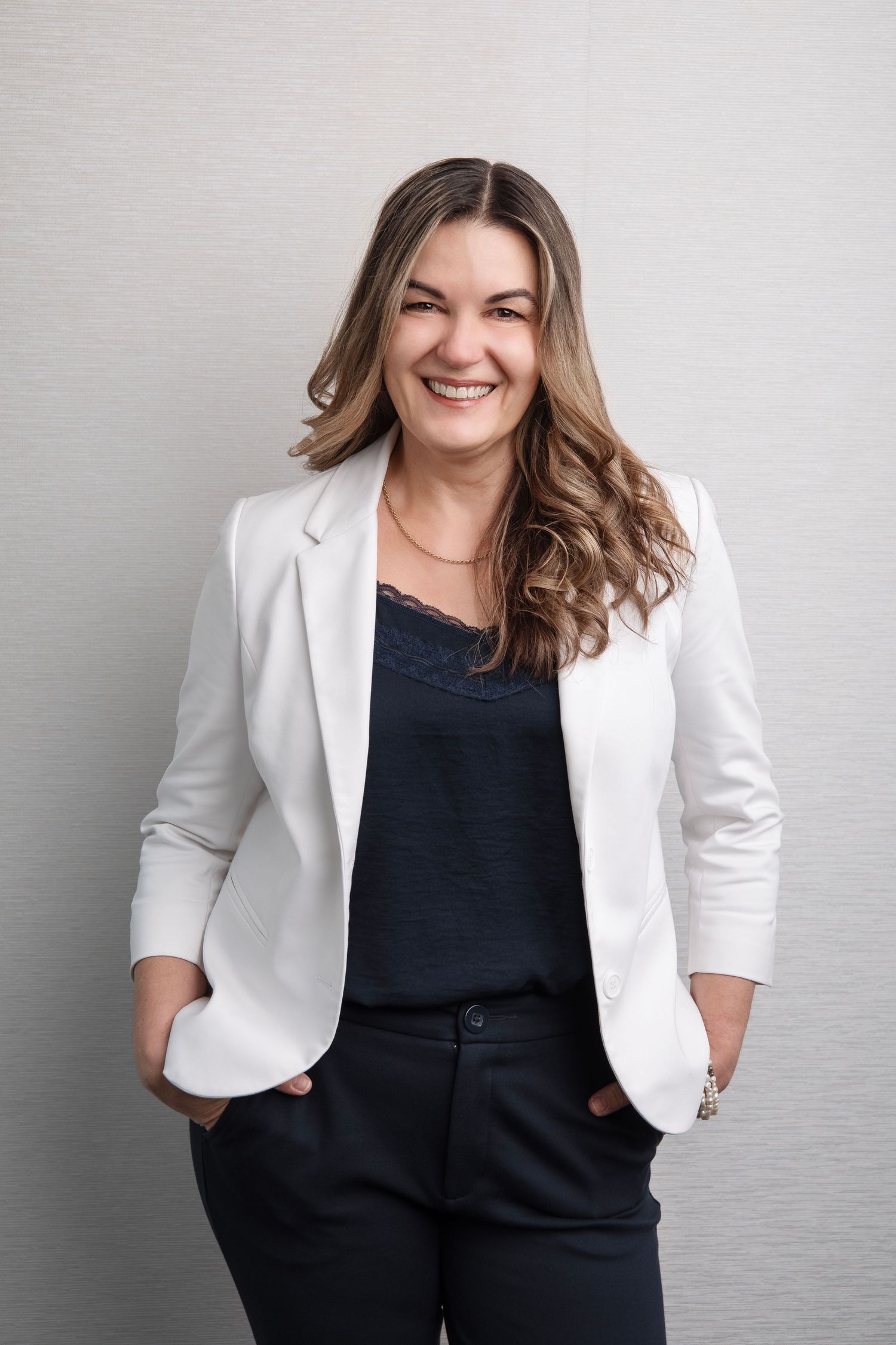 Professional portrait of someone in a crisp white blazer and black top standing confidently against a light gray background.