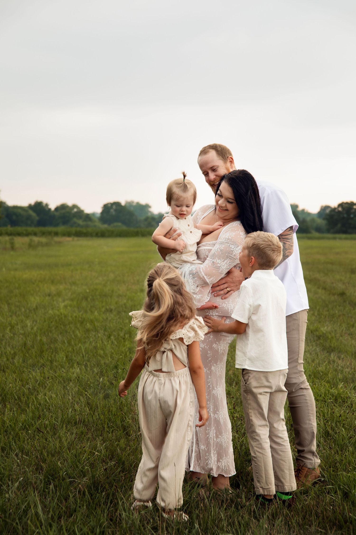Family dressed in white and beige embraces together while sharing joyful moments in a green field at sunset.