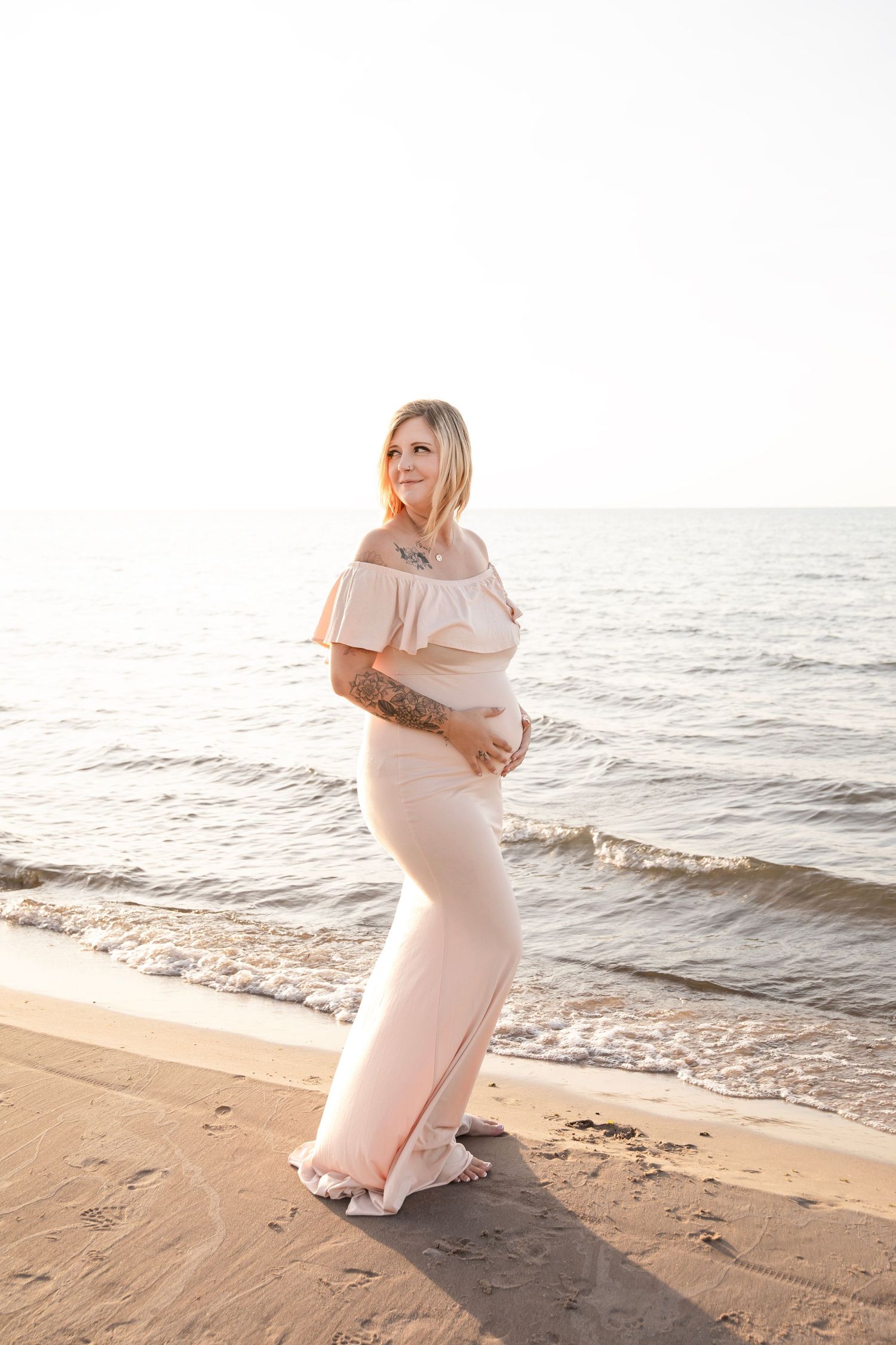 A pregnant person in a soft pink dress stands on a beach during sunset, captured in a series of four serene photos.