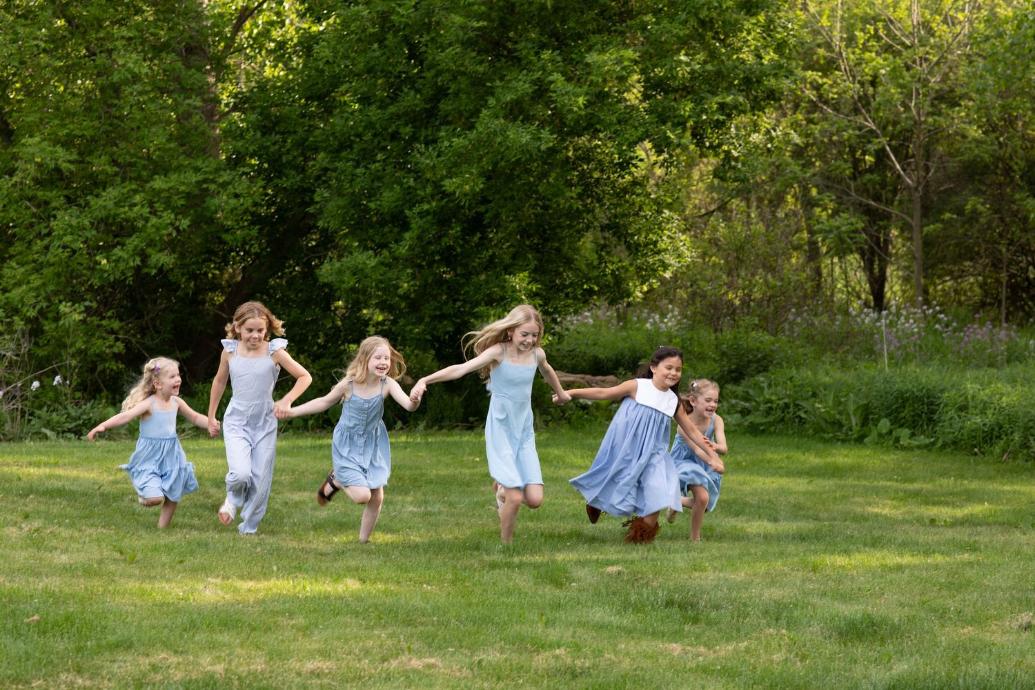 Six girls in blue dresses run hand-in-hand across a green lawn with lush trees in the background.