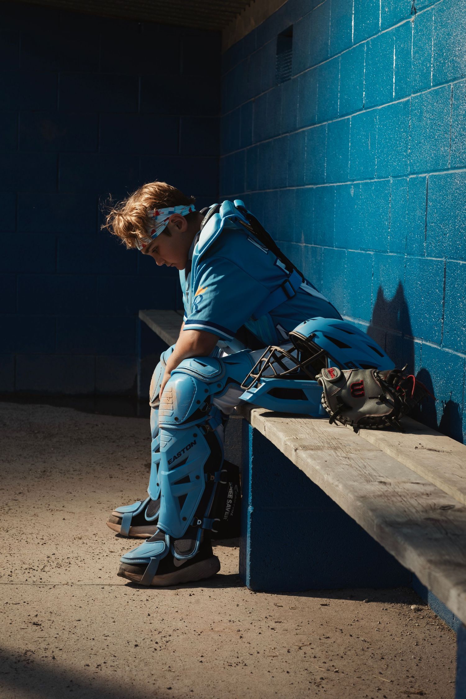 Young baseball catcher in full gear sitting alone on dugout bench, head down, wearing blue uniform and protective equipment.
