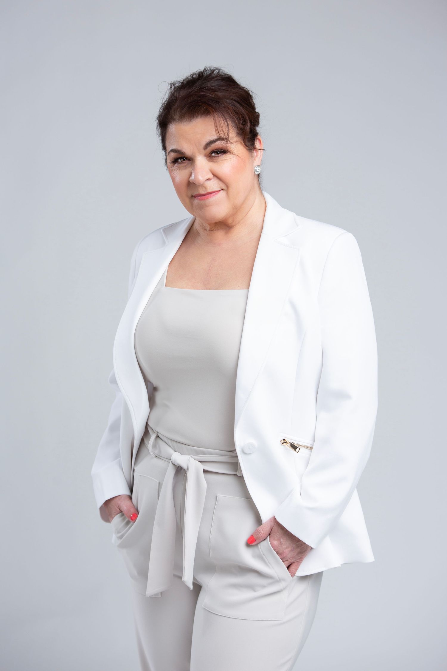 Professional studio portrait of a person in a white blazer and light-colored outfit posing with hands in pockets against grey backdrop.