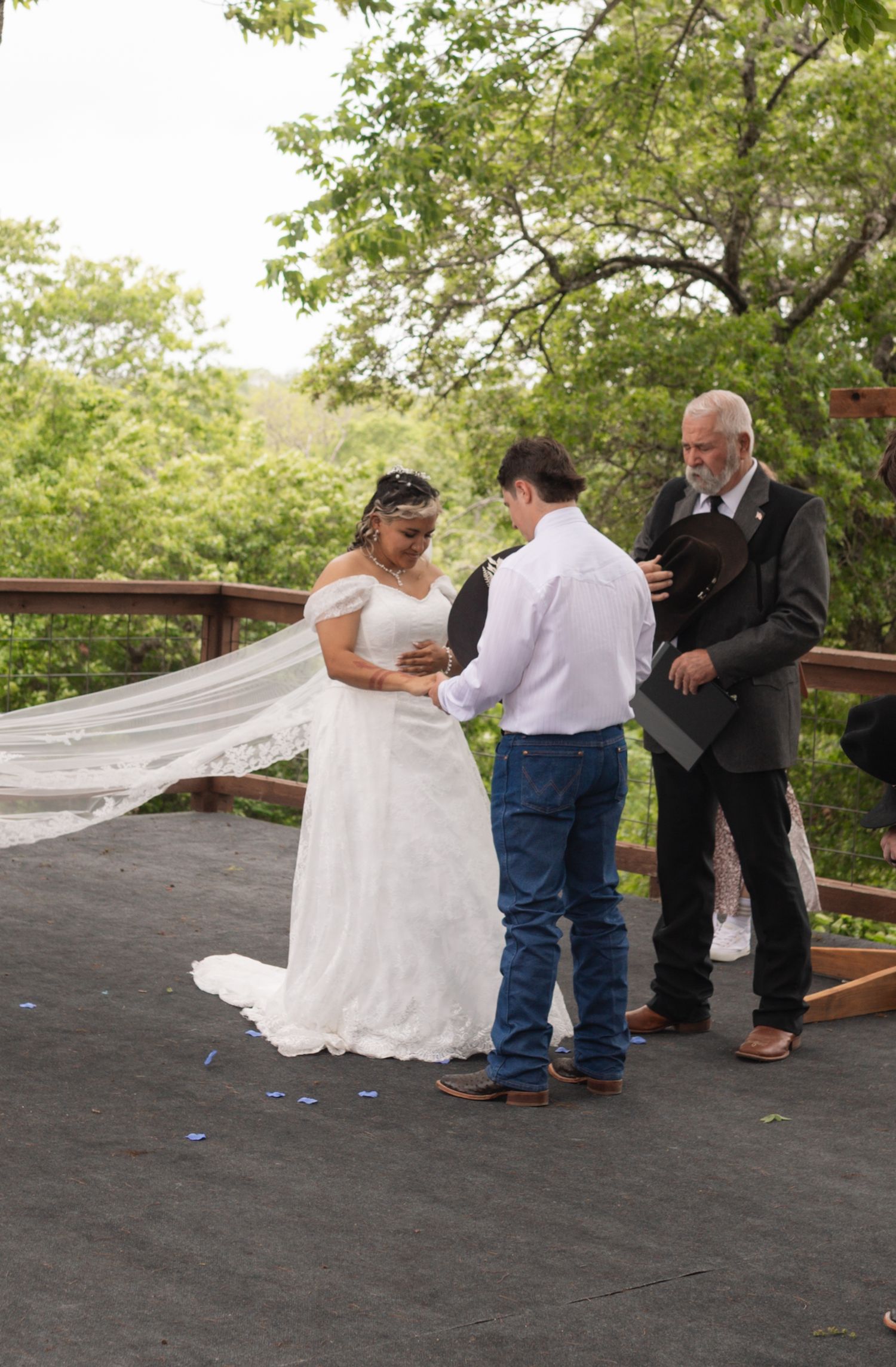 Outdoor wedding ceremony on a deck with bride in white gown and veil, groom in jeans, and officiant among green trees.