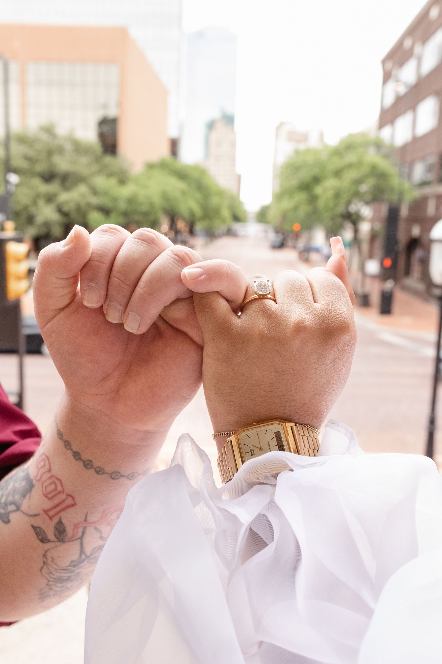 Close-up view of three hands with tattoos and rings doing a pinky promise gesture against a blurred urban background.