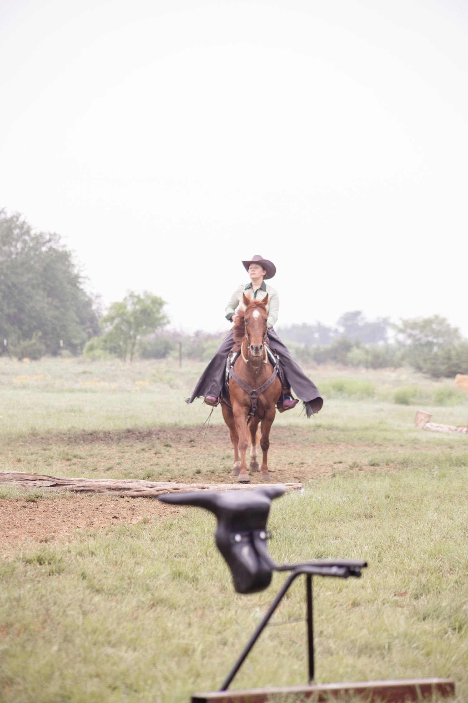 Cowboy riding a brown horse across a green field in misty conditions, with a gun target stand in the foreground.