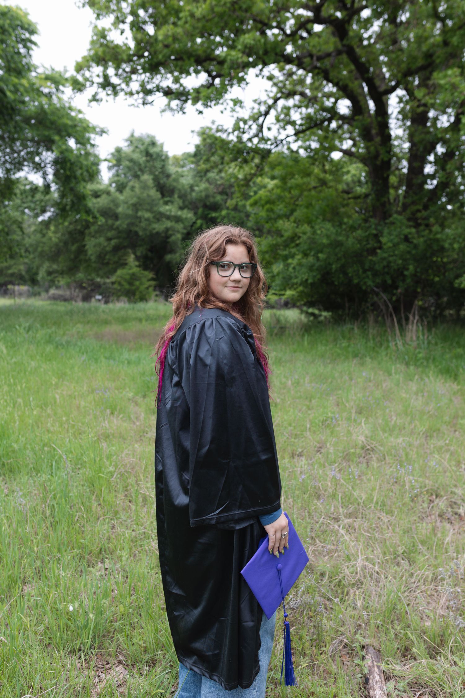 Young woman in black graduation gown and glasses holds blue cap in a green field with trees in background.