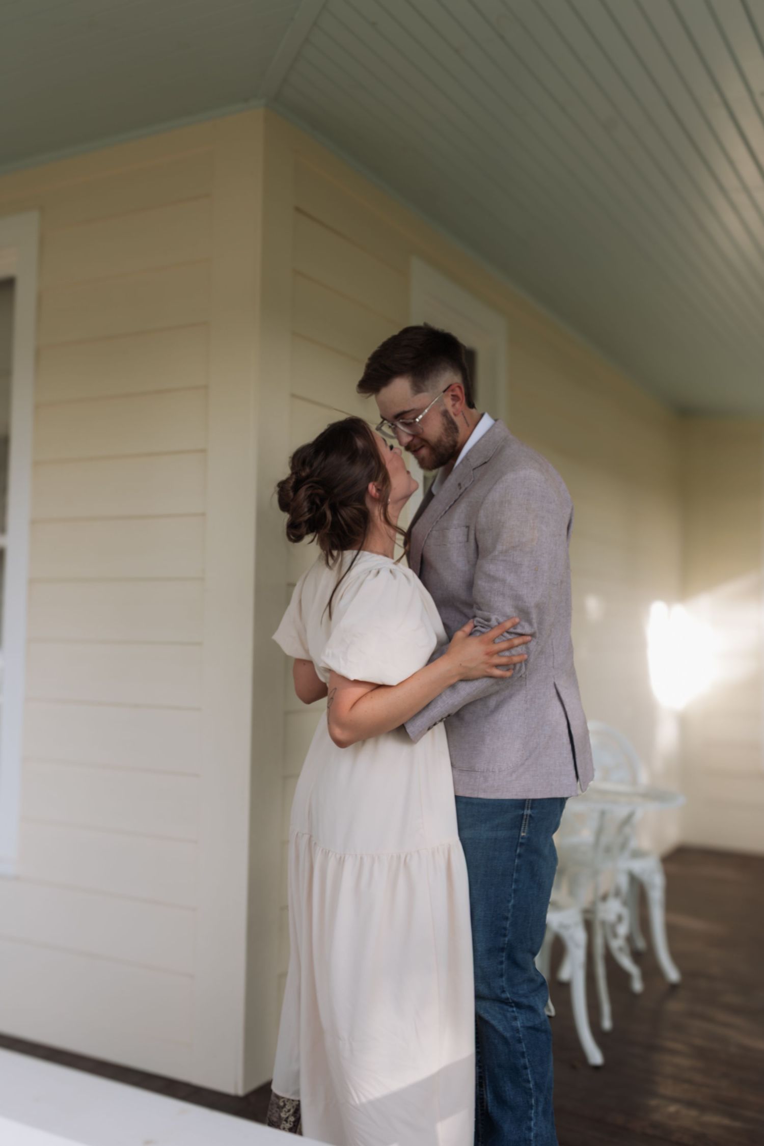 A couple embraces and nearly kisses on a sunlit porch, the man in a blazer and the woman in a white dress.