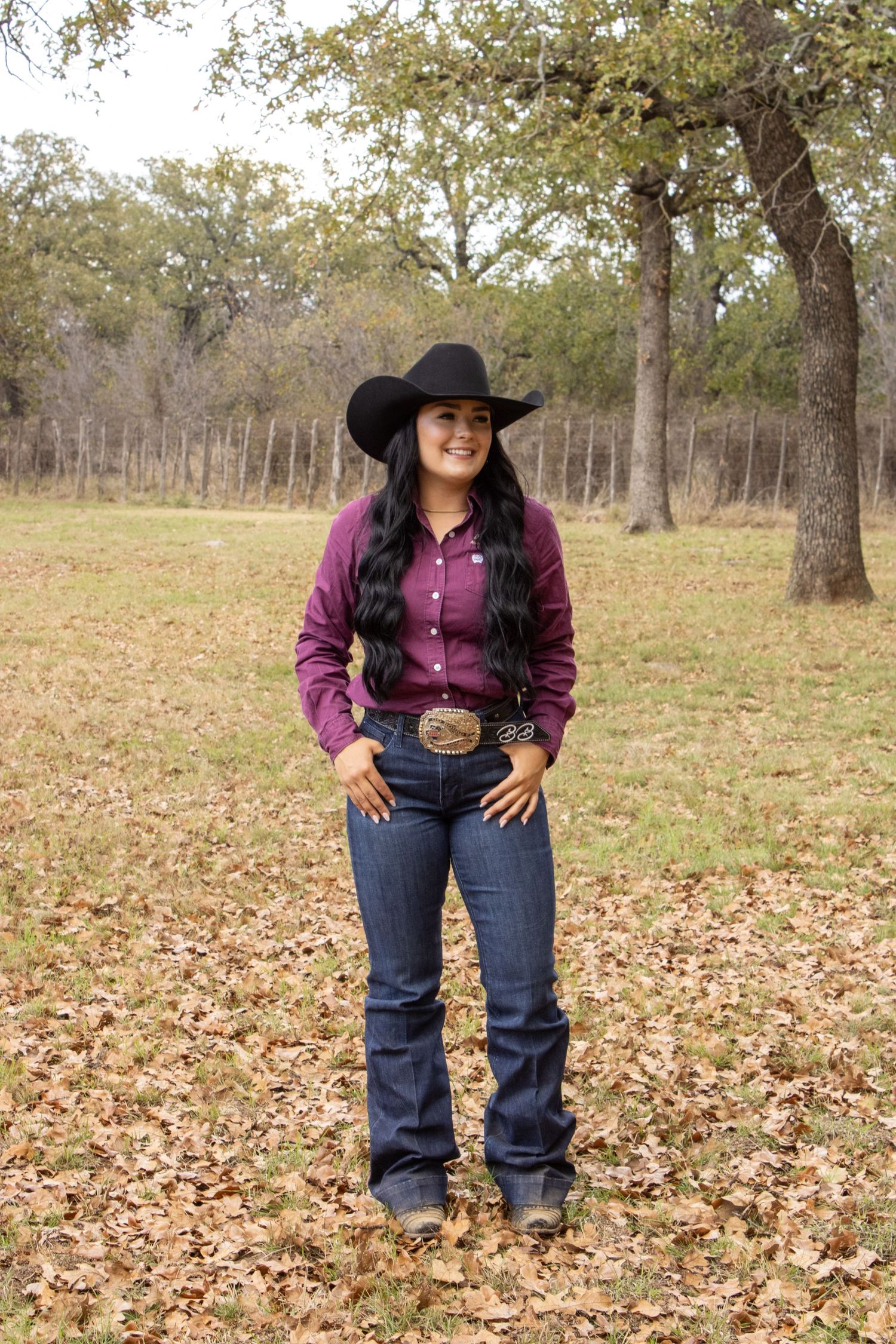 Western style outfit with purple button-down shirt, cowboy hat and jeans standing in autumn field with oak trees.