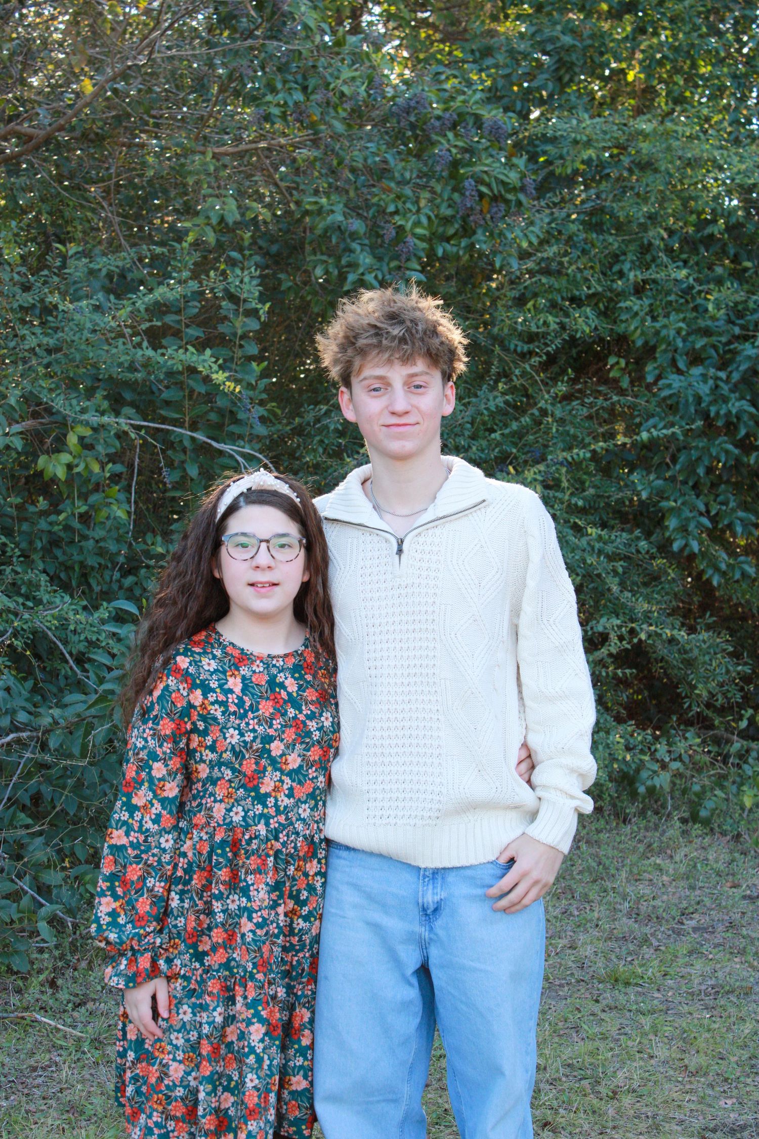 Two people stand together outdoors in front of green foliage wearing casual clothing on a sunny day.