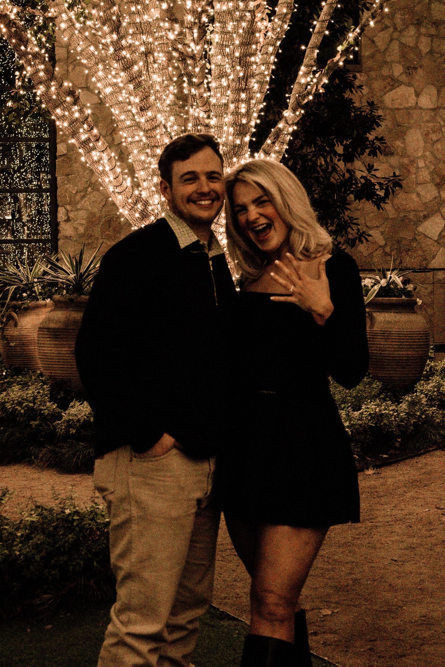 A couple shares a joyful moment together in front of a wall decorated with twinkling string lights.