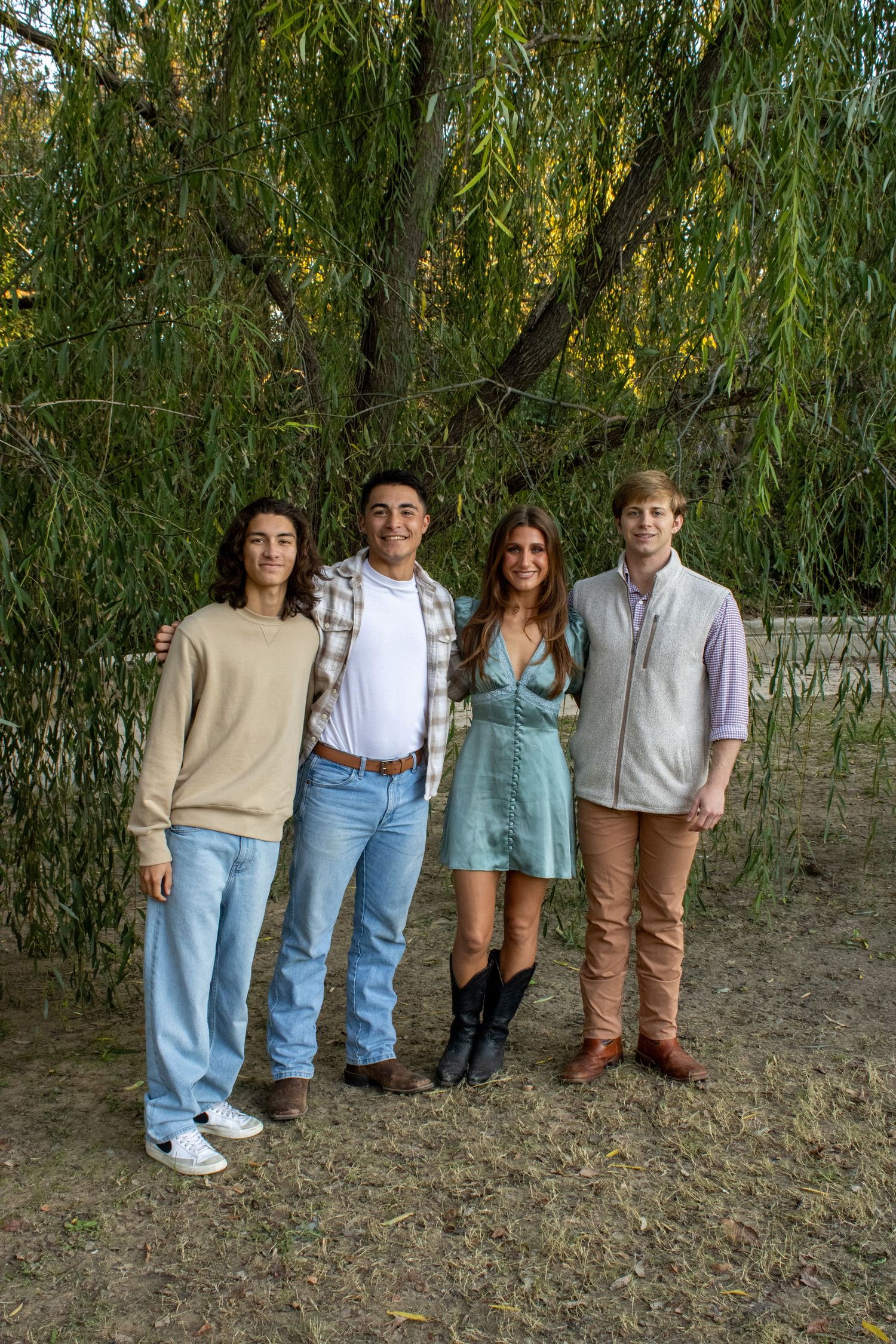 A group of friends in casual outfits stand together against a backdrop of drooping willow tree branches.
