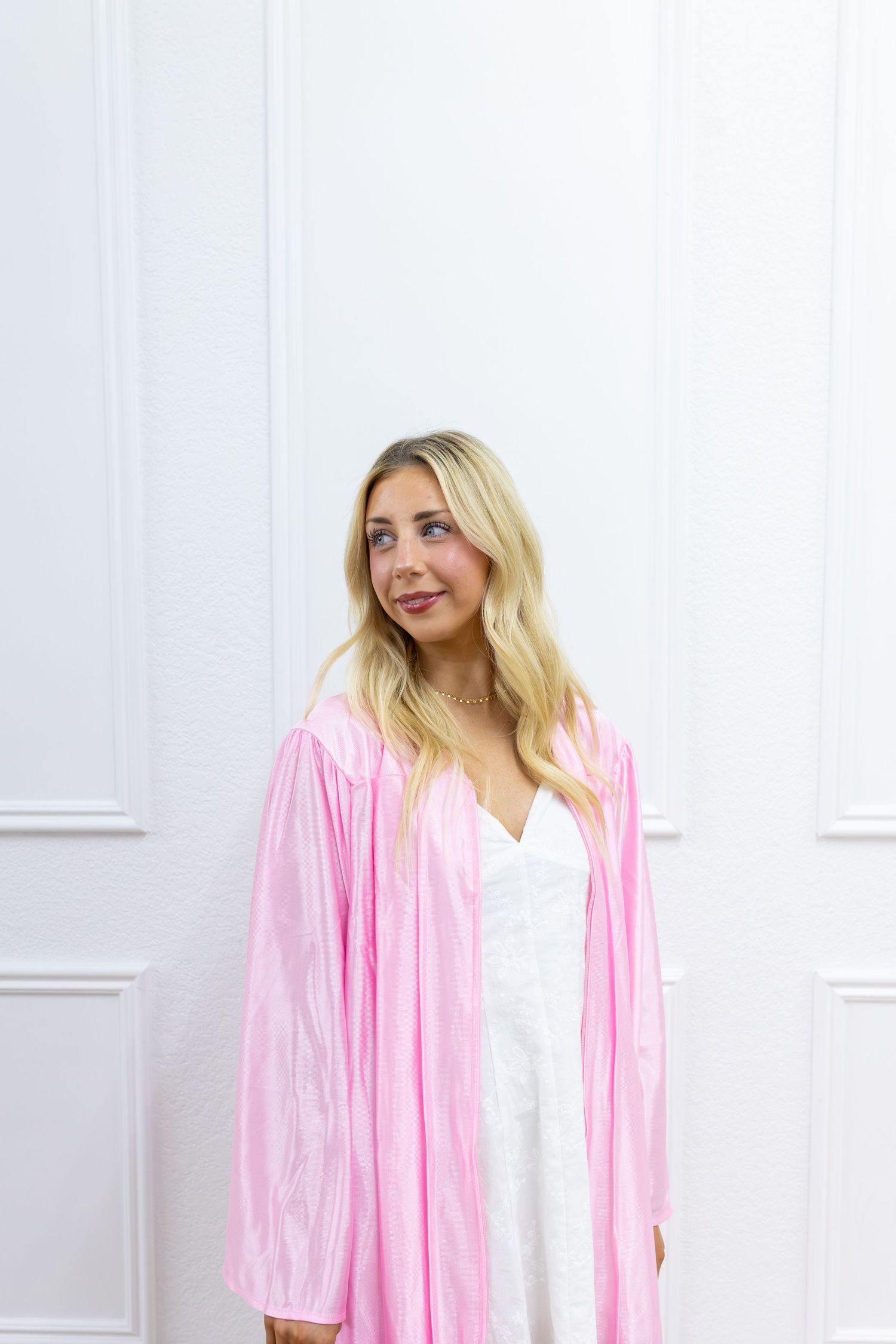 Blonde woman wearing a pink graduation gown over a white dress, posing against a white paneled wall.