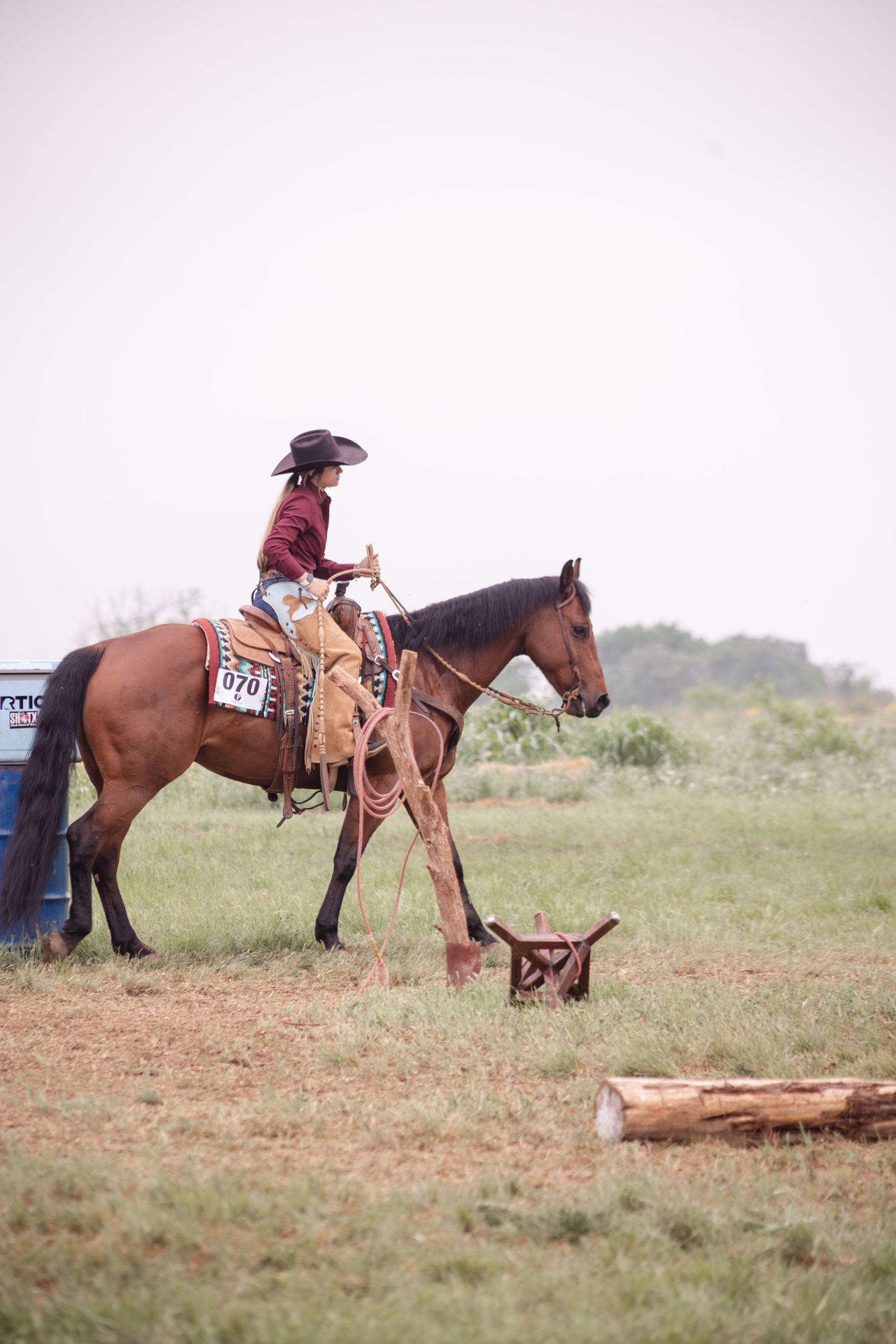 Cowgirl in hat rides brown horse at rodeo competition, wearing number 070, with barrels visible in grassy field.