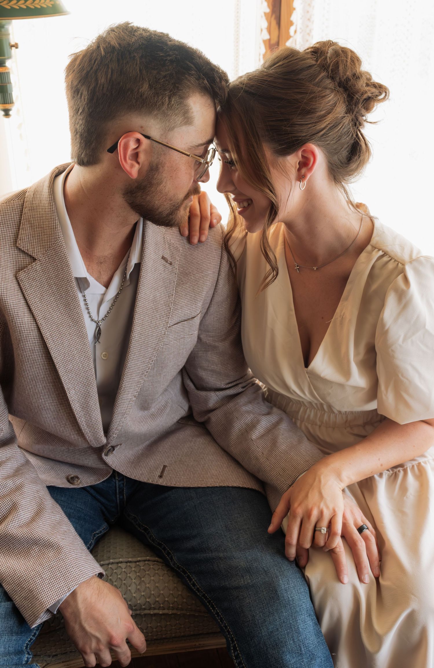 A couple in elegant attire sits close together, smiling and sharing an intimate moment indoors.