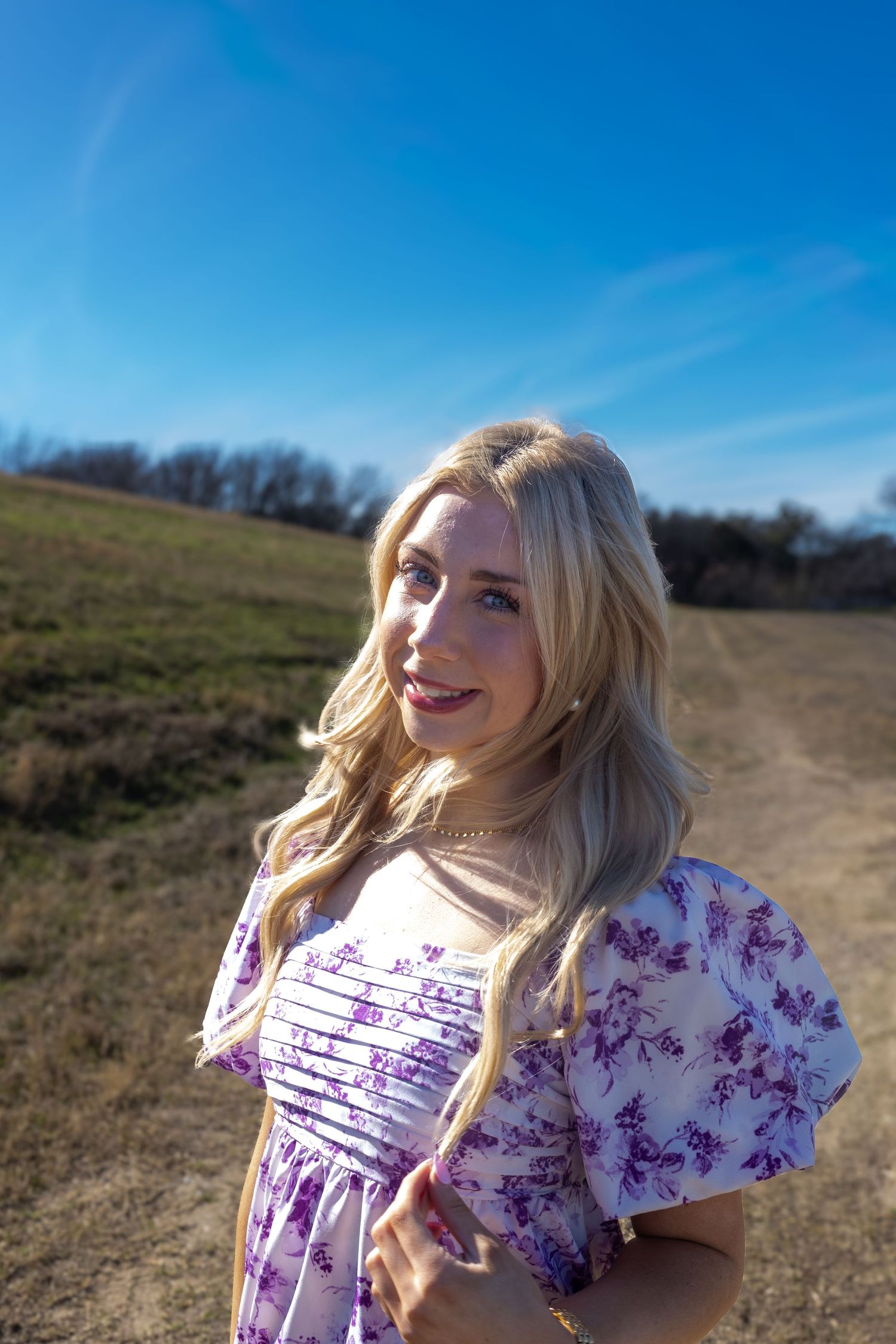 A person in a floral dress poses in a sunny field with blue sky and bare trees in the background.
