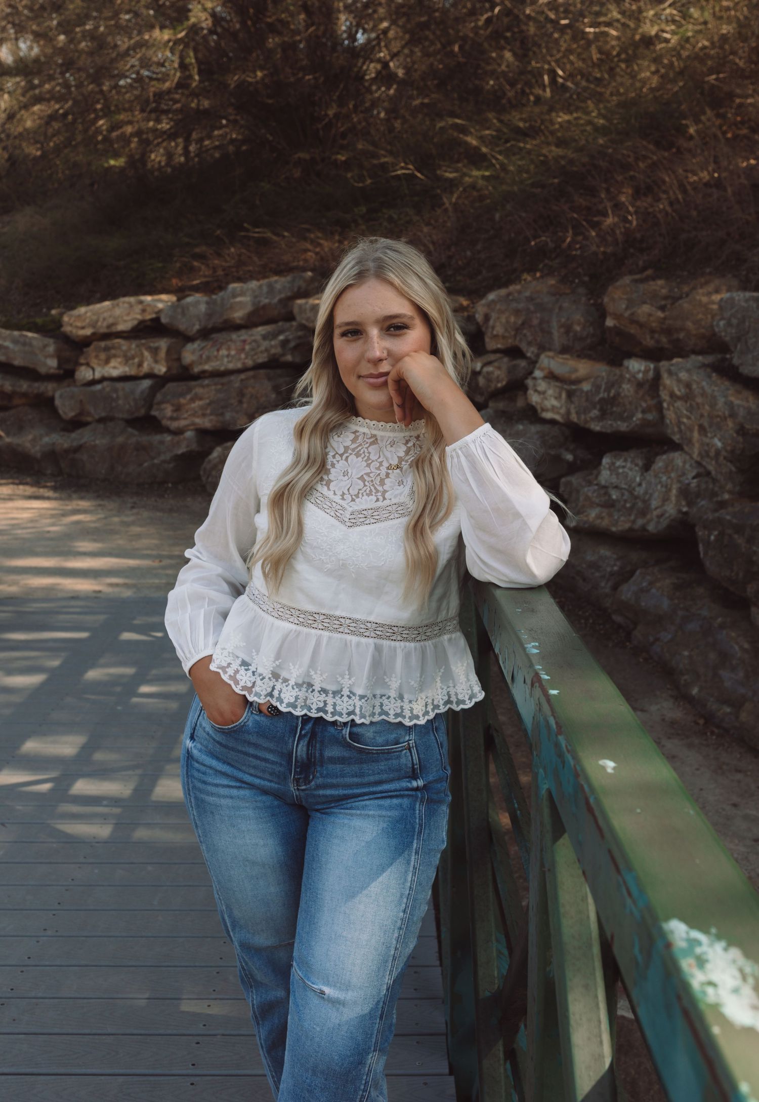 A person wearing a white lace top and jeans leans against a bridge railing in a natural outdoor setting.