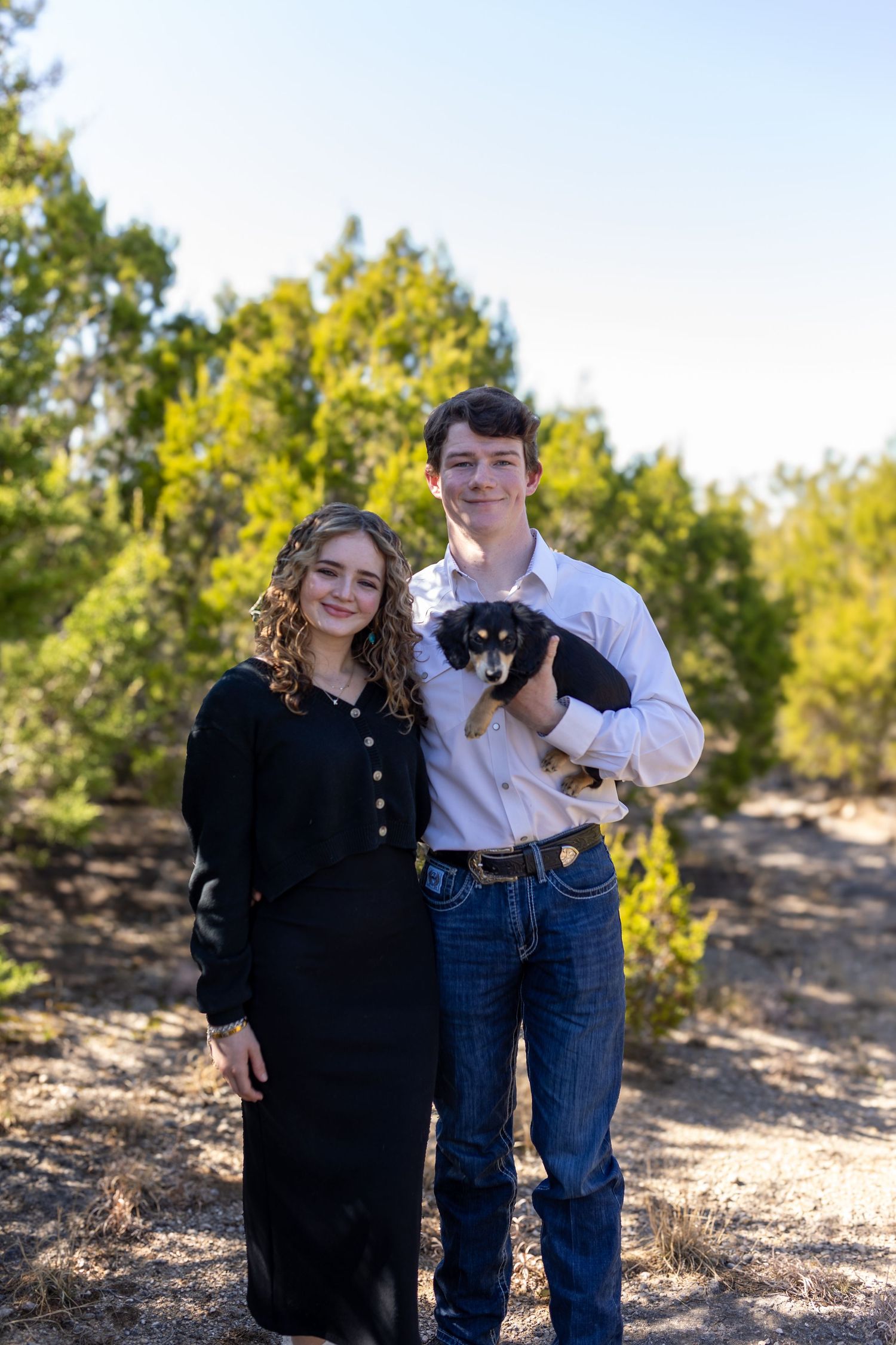 A couple poses outdoors with their dog on a sunny day among trees and natural terrain.