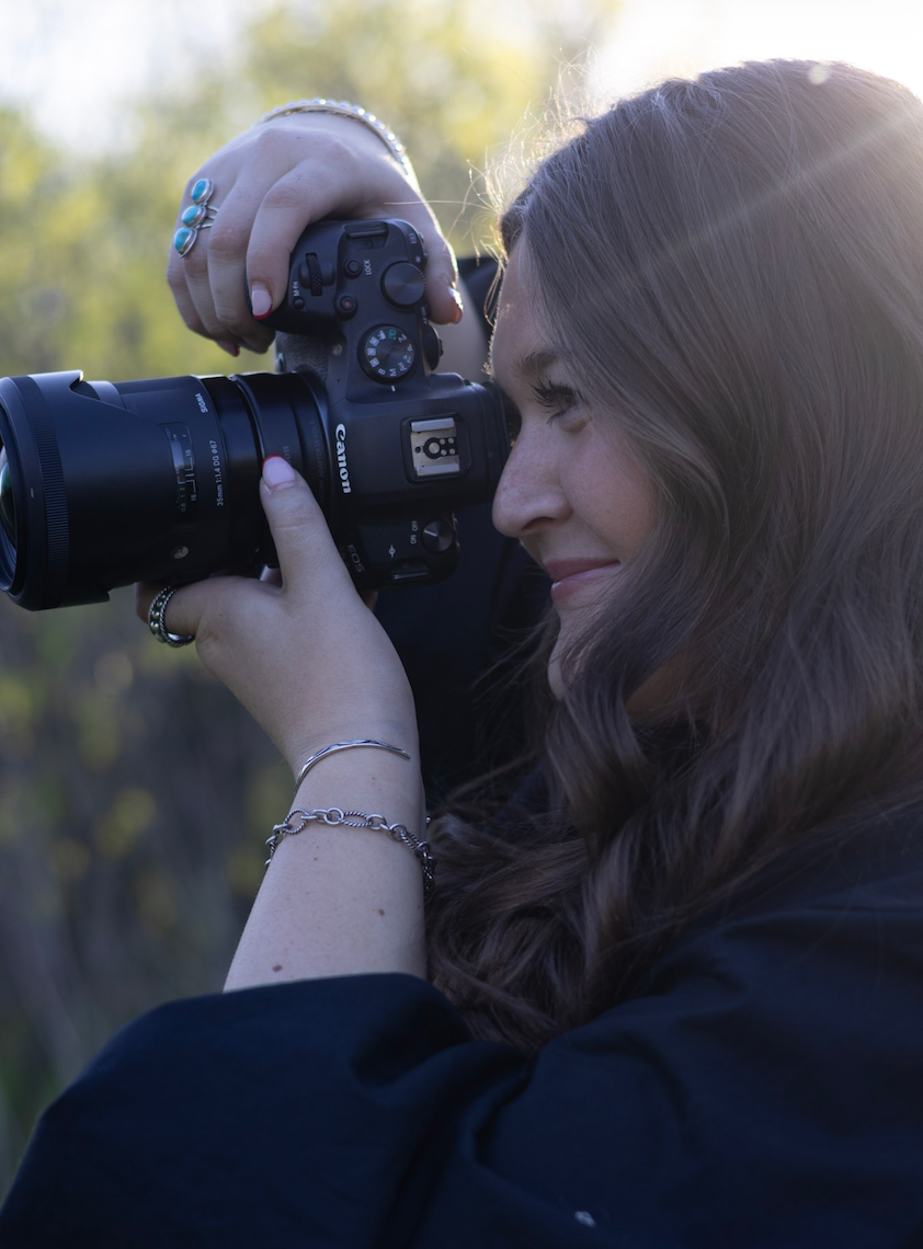 A photographer in a dark shirt uses a DSLR camera to take photos outdoors with natural lighting in the background.