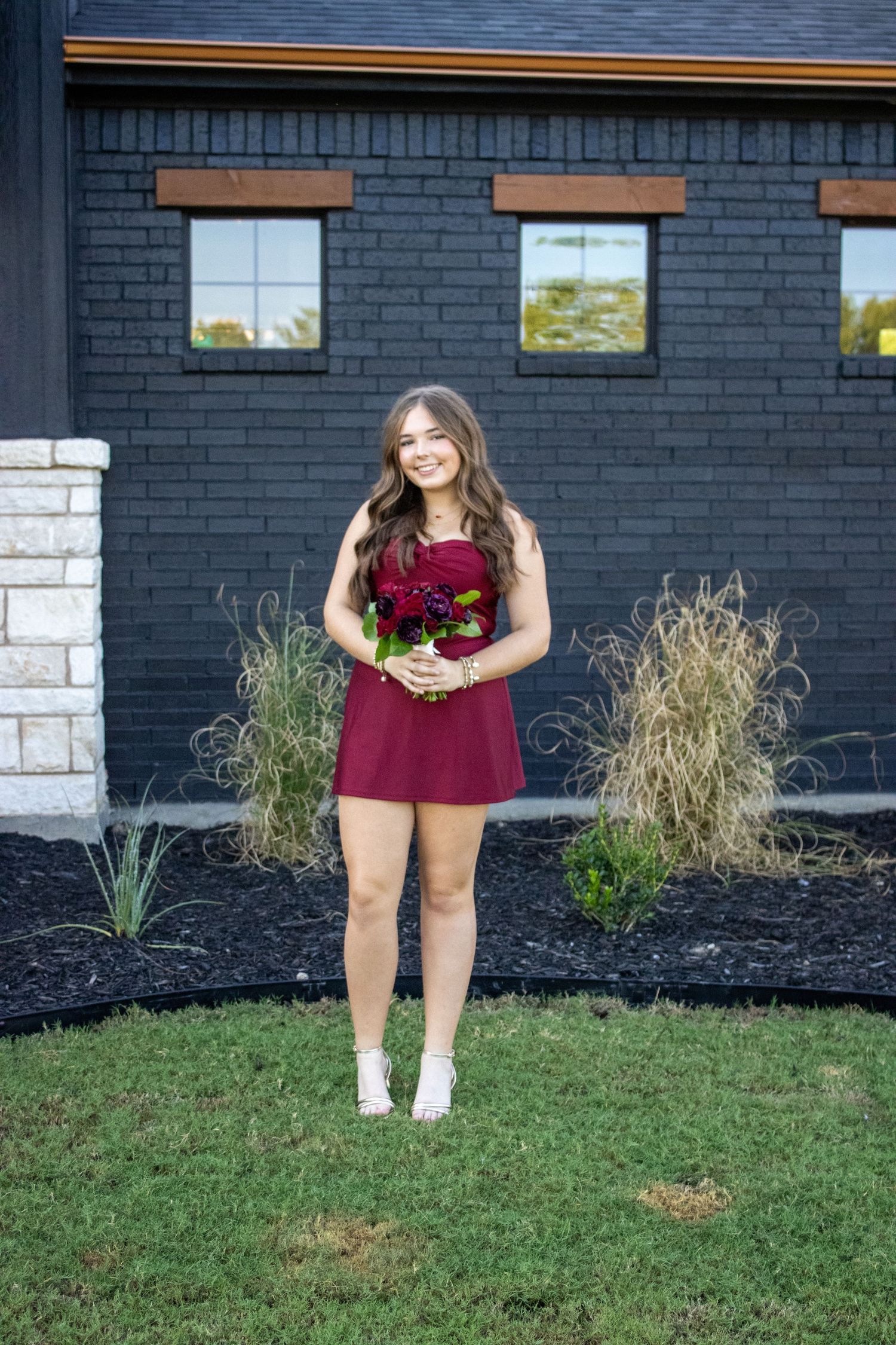 Portrait session against dark brick building with decorative landscaping.