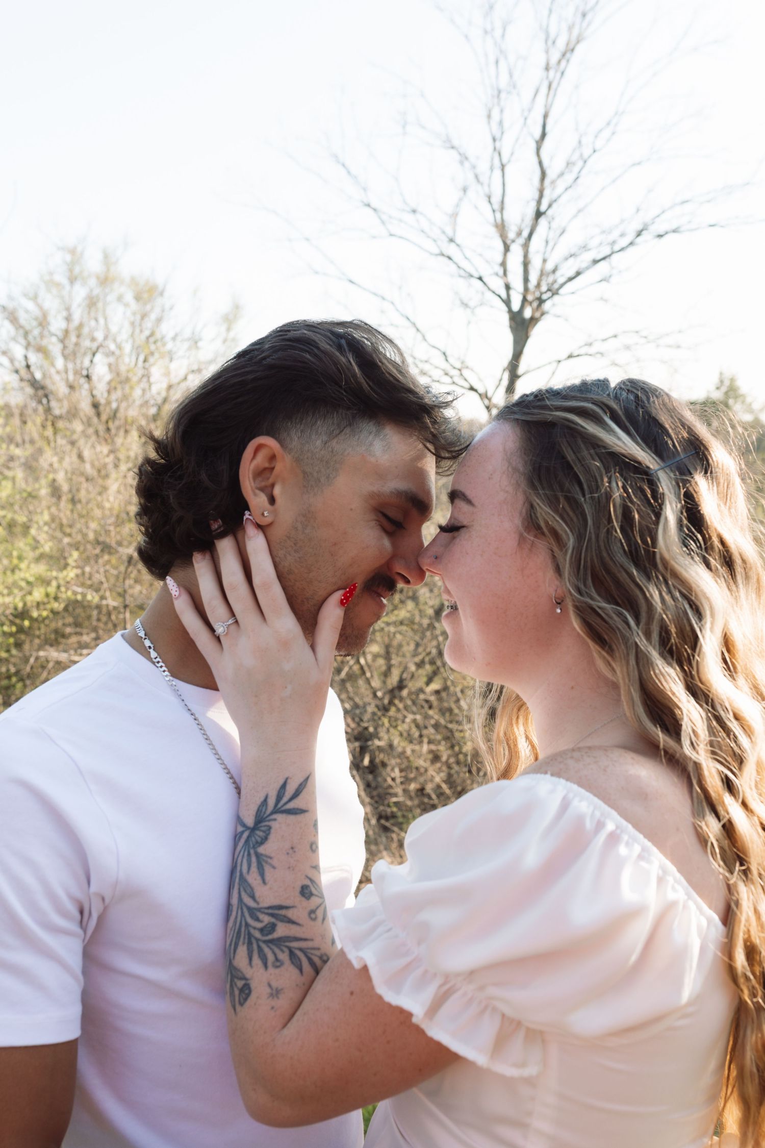 Couple sharing a romantic moment outdoors, man with tattoos and woman in white dress, noses touching, smiling, trees in background.