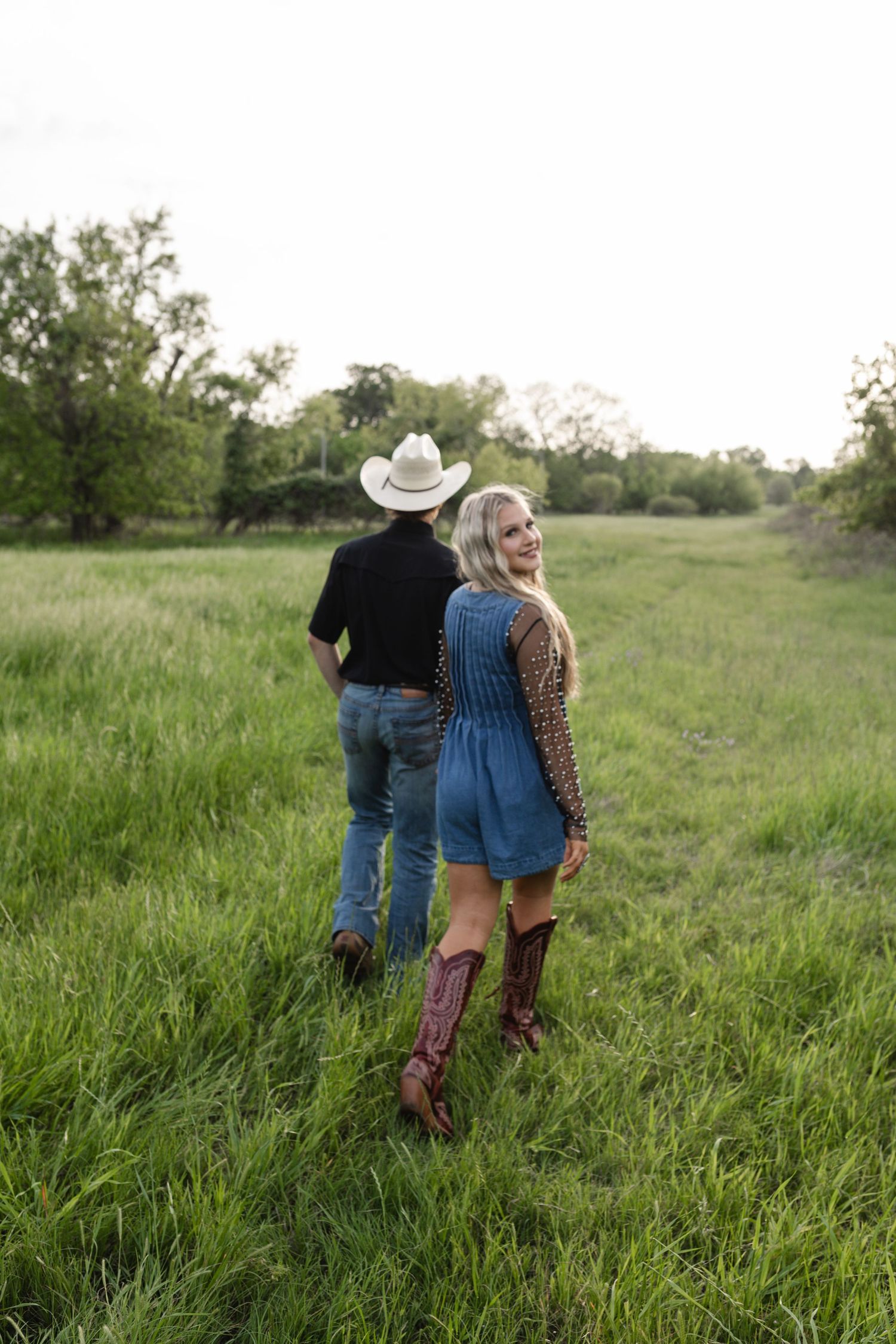 Couple walking in green field, man in cowboy hat and woman in denim romper with cowboy boots.