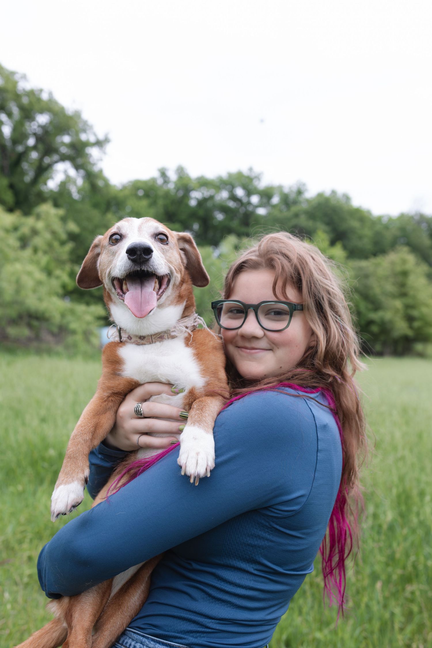 A smiling girl with glasses holds a happy brown and white dog in a lush green outdoor field.