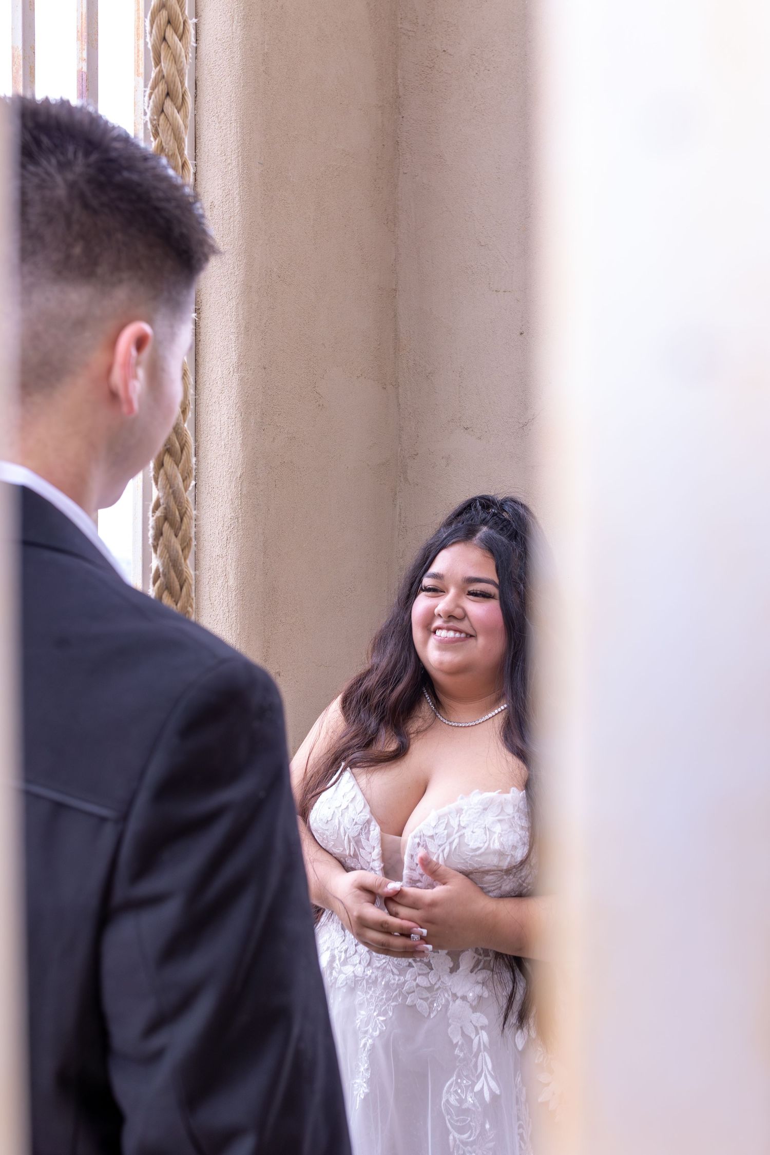 Bride in lace wedding gown smiling at groom in black suit during romantic first look ceremony.
